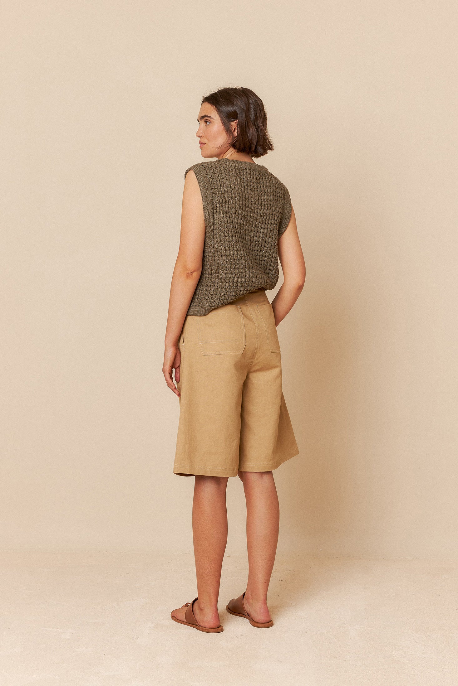 Woman wearing a sleeveless green top and beige bermuda shorts against a beige background