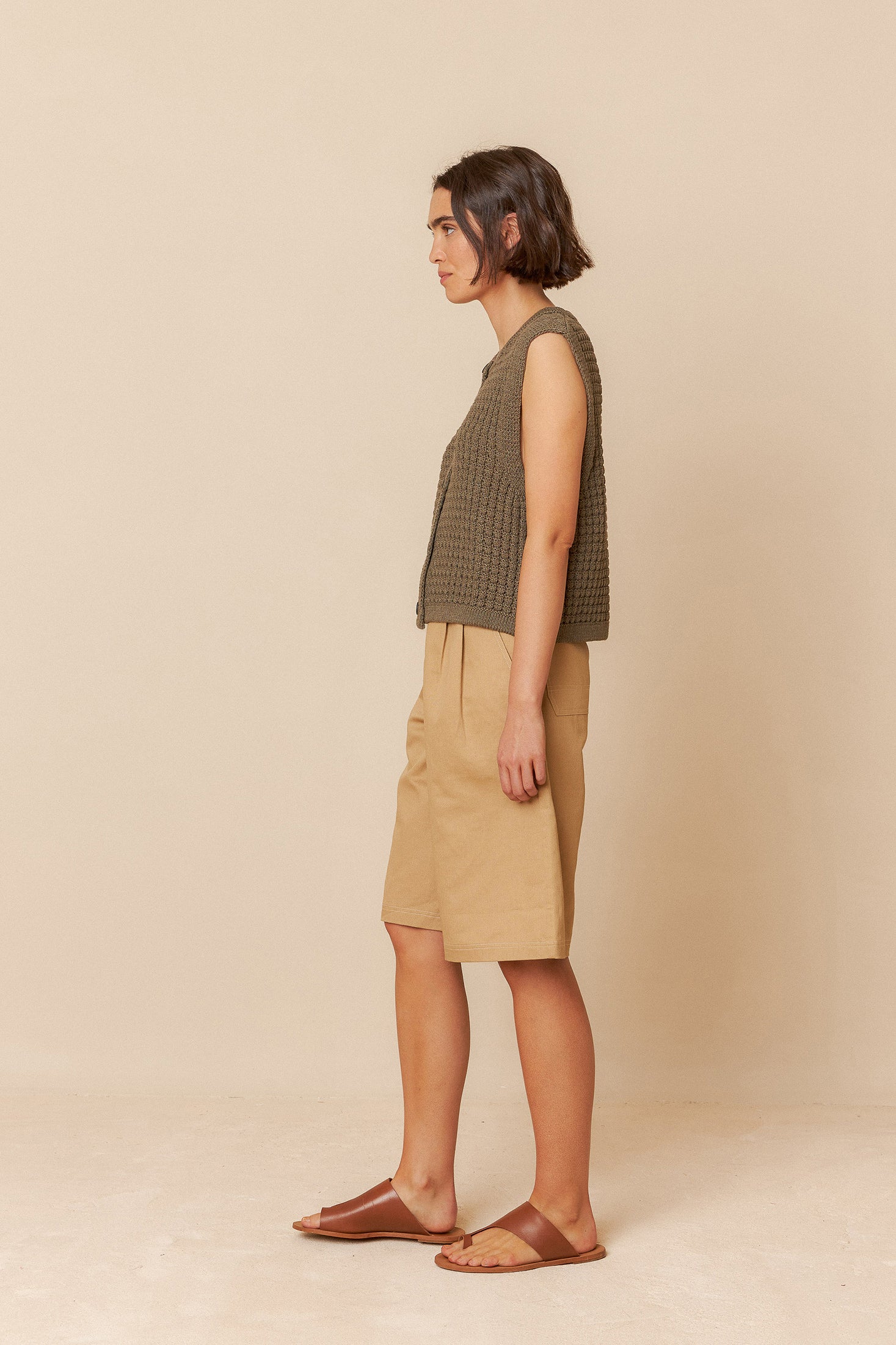 Woman wearing a sleeveless green top and beige bermuda shorts against a beige background