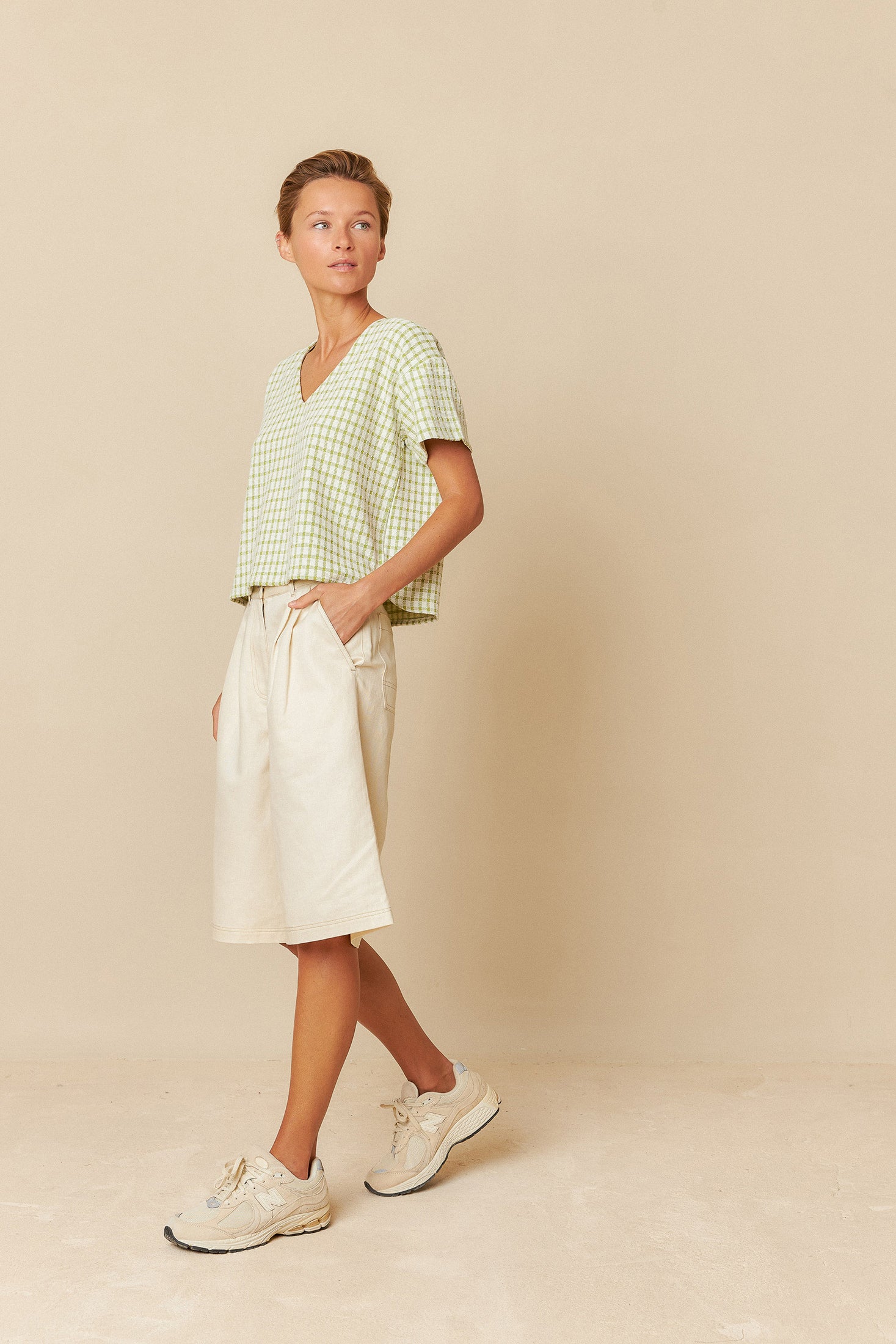 Woman wearing a green and white top  and ecru bemuda shorts standing in front of a colourful wall.