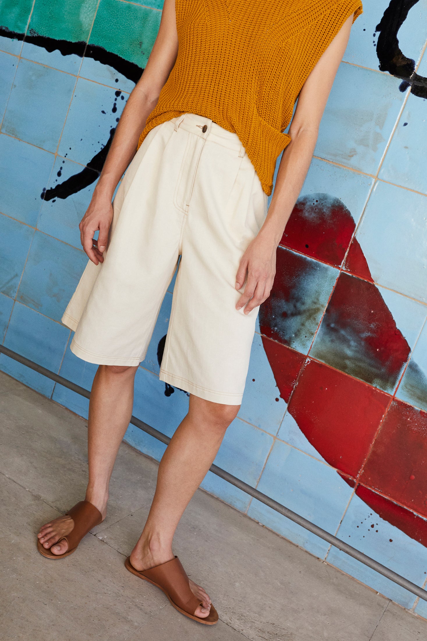 Woman wearing a mustard sleeveless sweater and ecru bemuda shorts standing in front of a colourful wall.