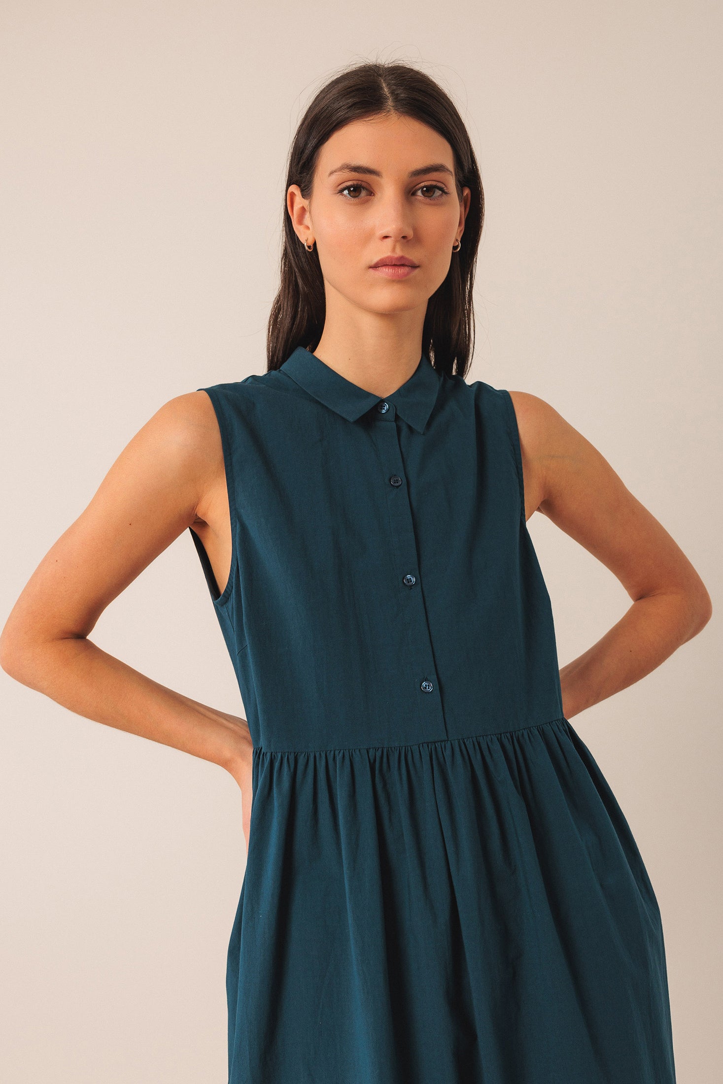 Woman wearing a teal sleeveless shirt dress against a plain background