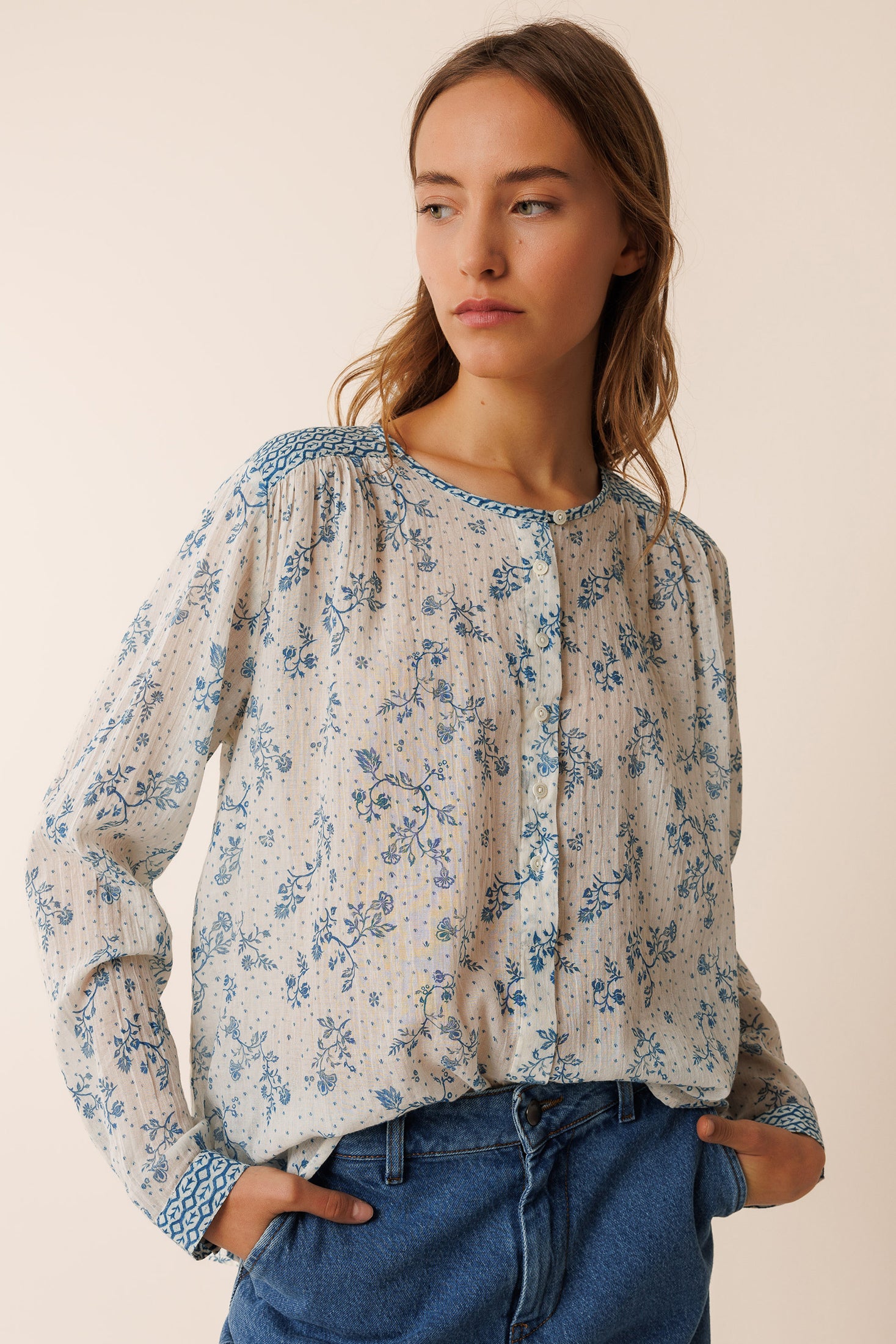 Woman wearing a floral long sleeve cotton blouse and blue jeans against a beige wall