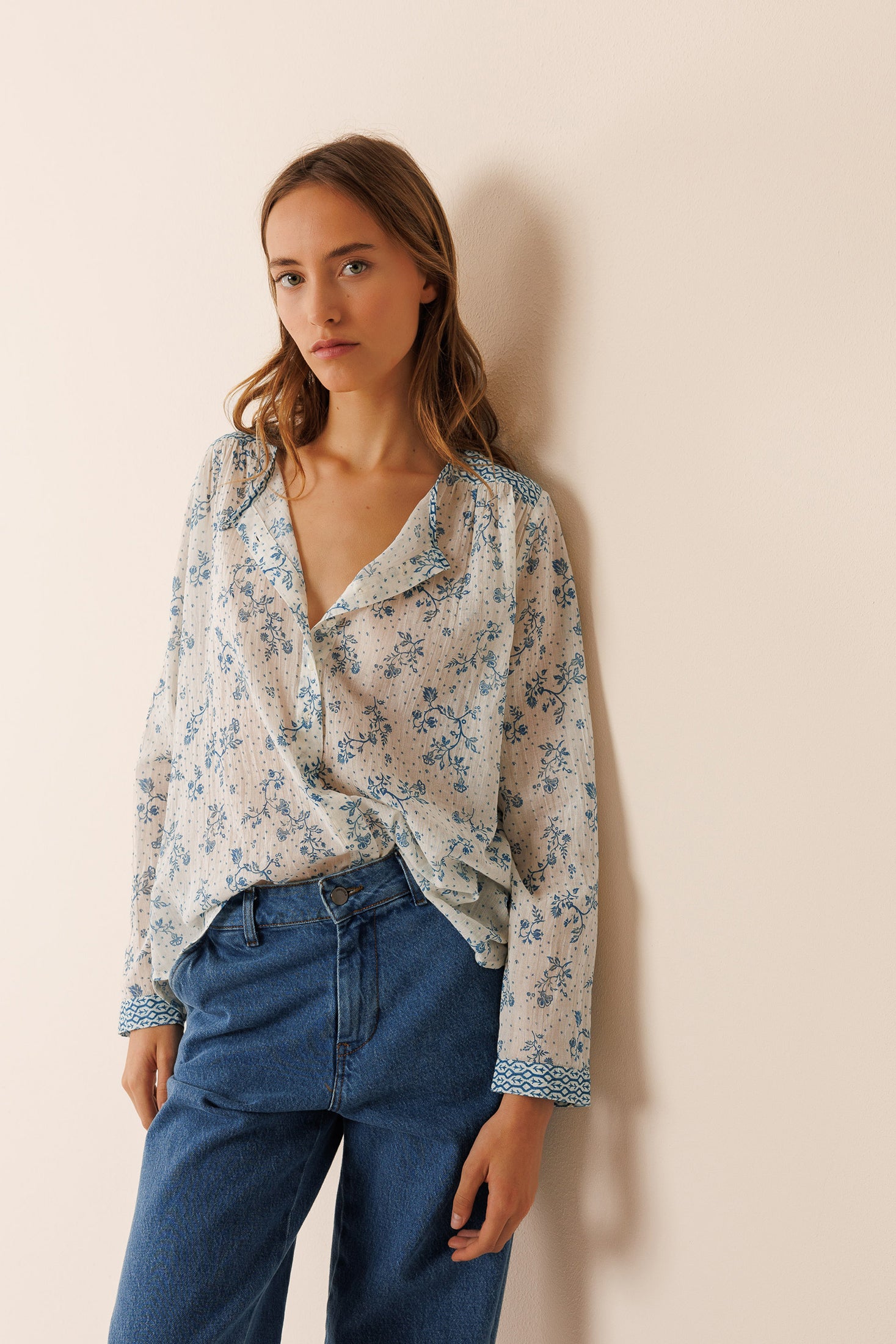 Woman wearing a floral long sleeve cotton blouse and blue jeans against a beige wall