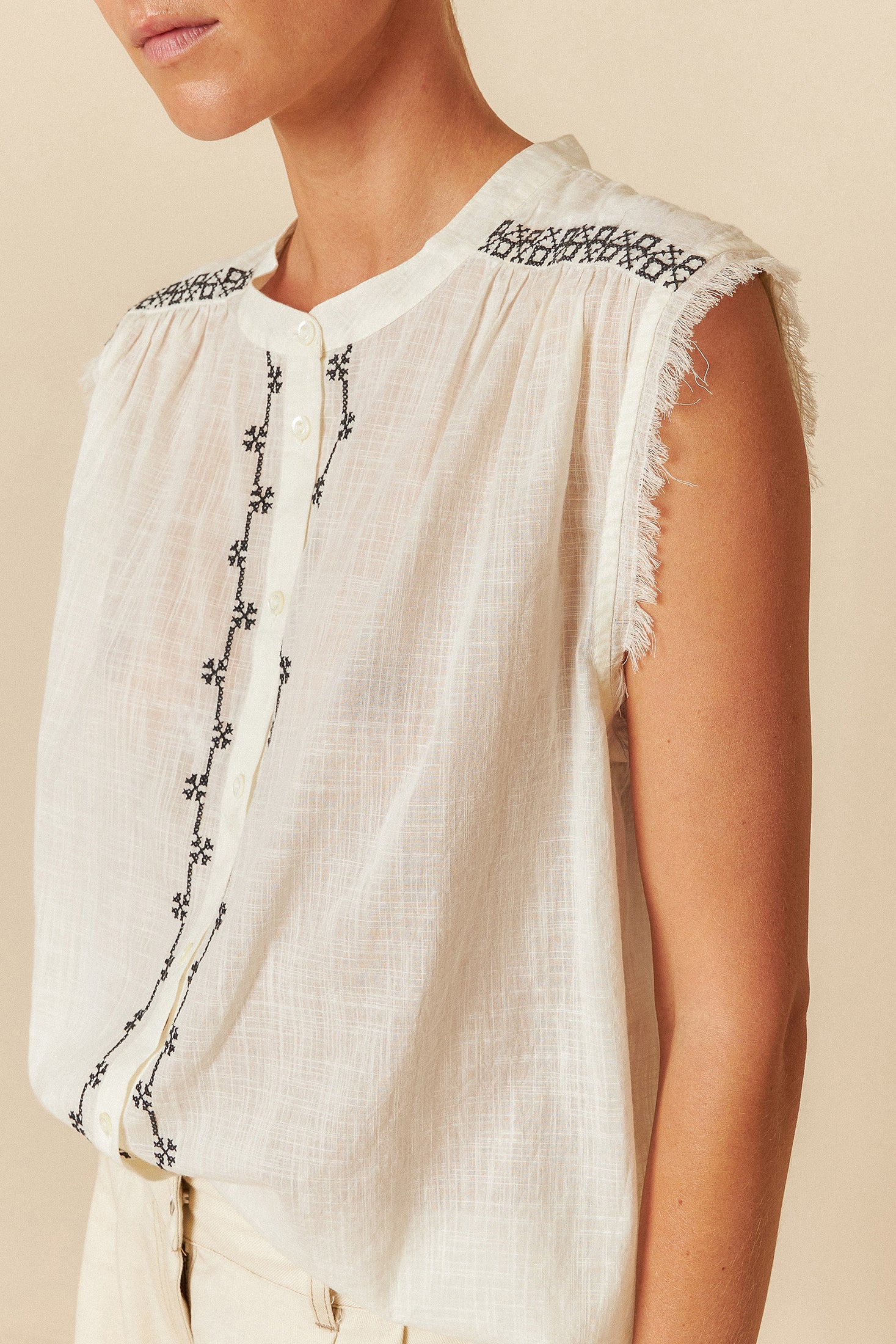 White sleeveless top with black embroidery and fringes on a beige background