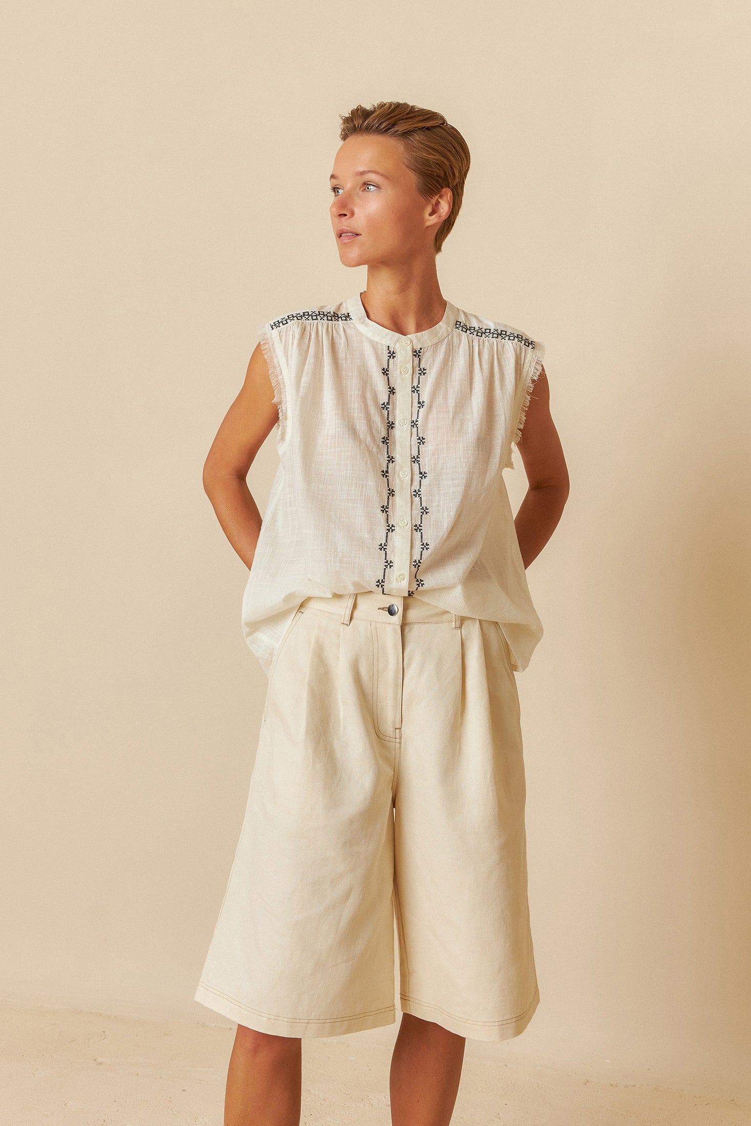 Woman wearing a white sleeveless top with black embroidery and fringes on a beige background with white shorts. 