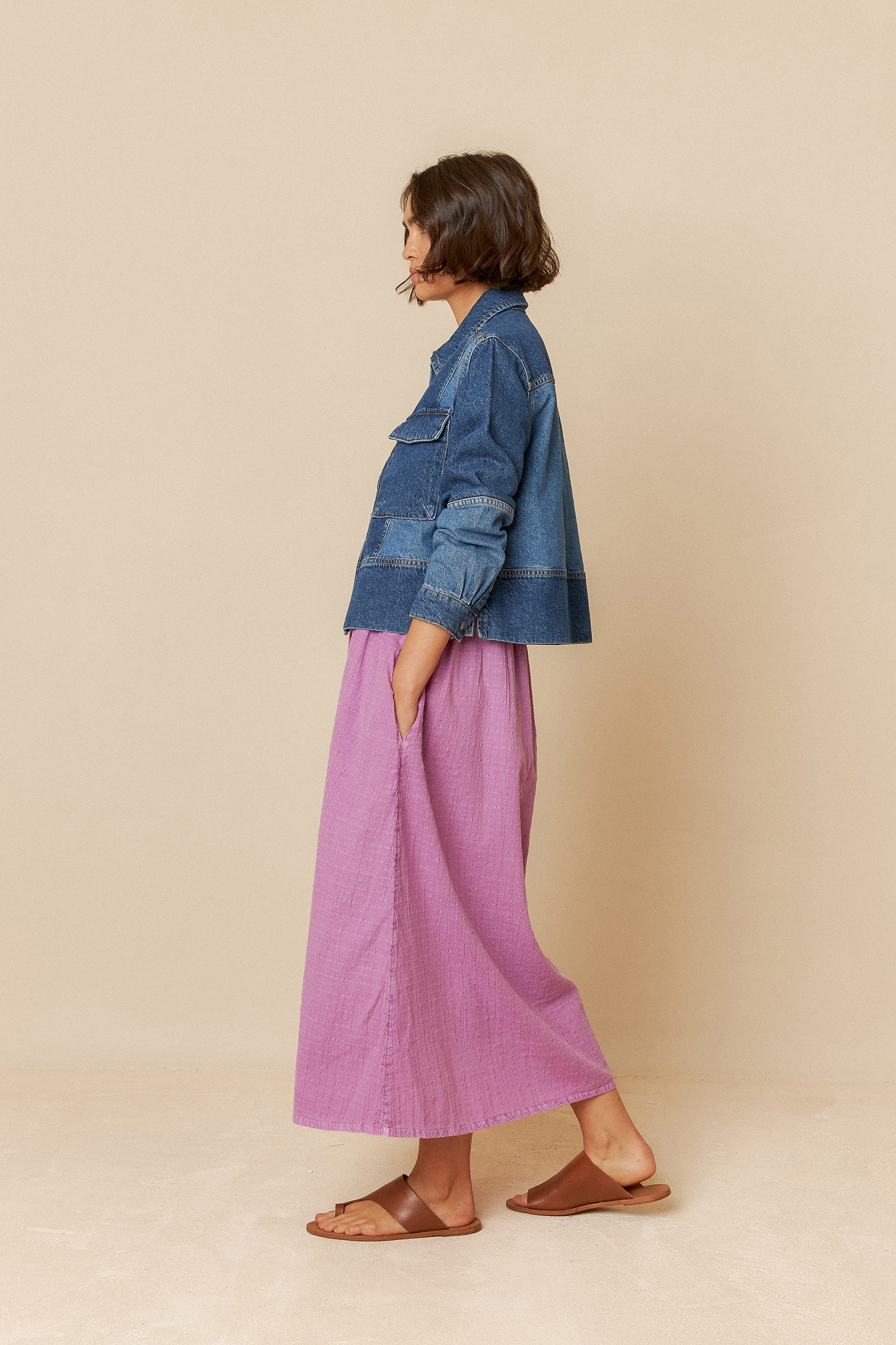 Model wearing a blue denim jacket with silver domes and multi coloured patchwork over a pink dress on a beige background