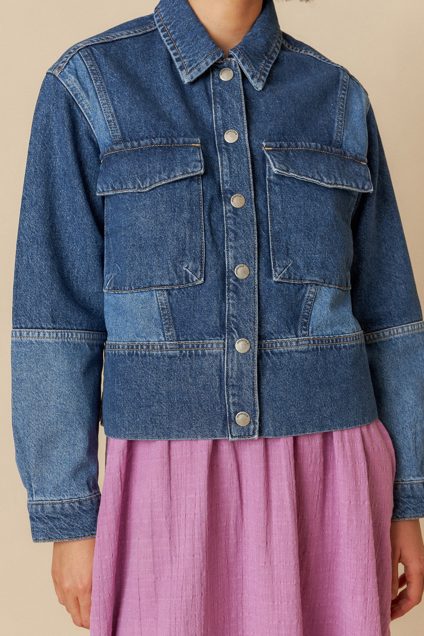 Model wearing a blue denim jacket with silver domes and multi coloured patchwork over a pink dress on a beige background