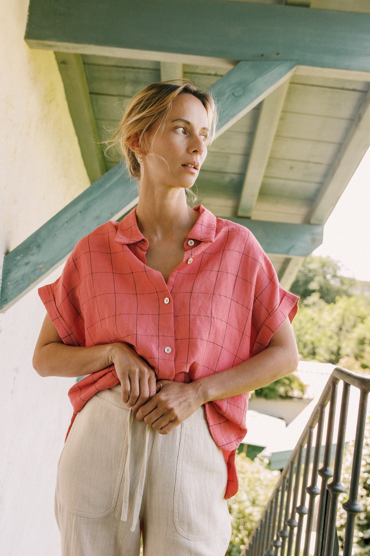 Woman wearing a pink checkered shirt and beige pants standing on a balcony.