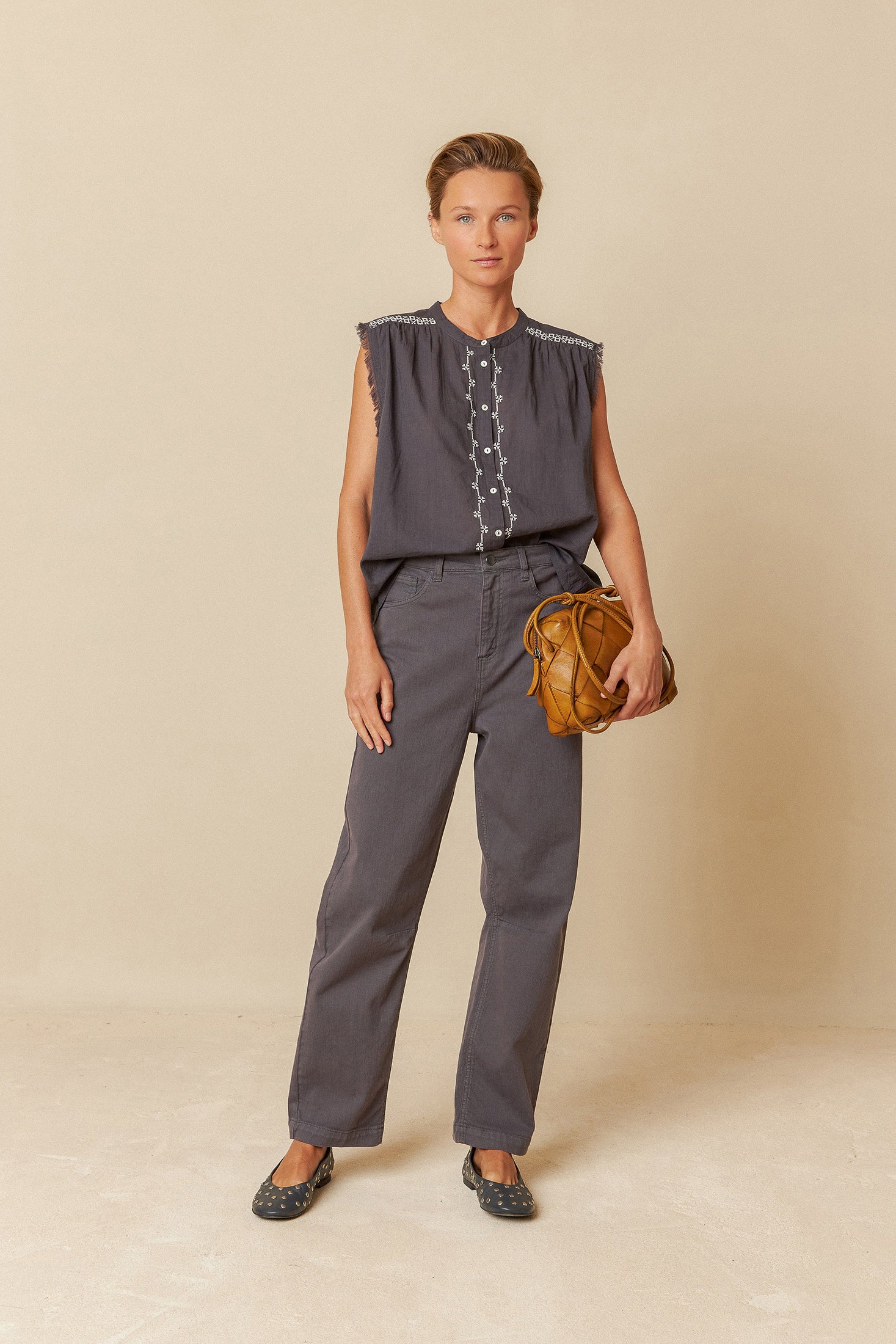 Woman wearing a black sleeveless top with white embroidery and black pants on a light grey background
