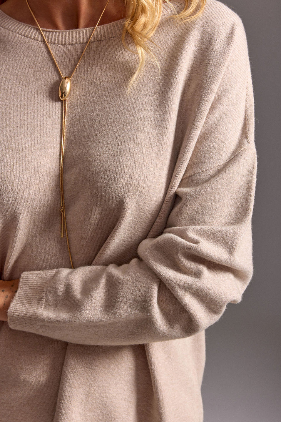 Person wearing a beige sweater with a gold necklace against a neutral background