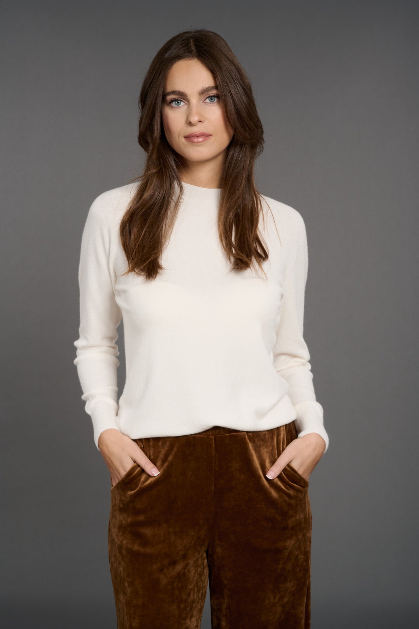 Woman wearing a white sweater and brown pants against a gray background