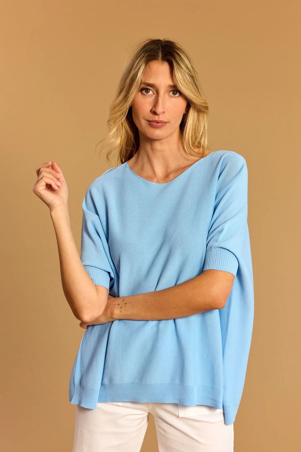 Women wearing light blue short sleeve knit poncho sweater with white jeans on a tan background. 
