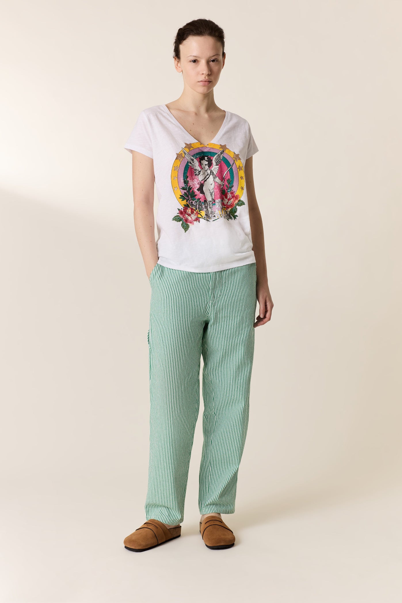 A woman stands against a plain beige studio background wearing a white graphic V-neck T-shirt, mint green wide-leg pants, and brown slip-on shoes.