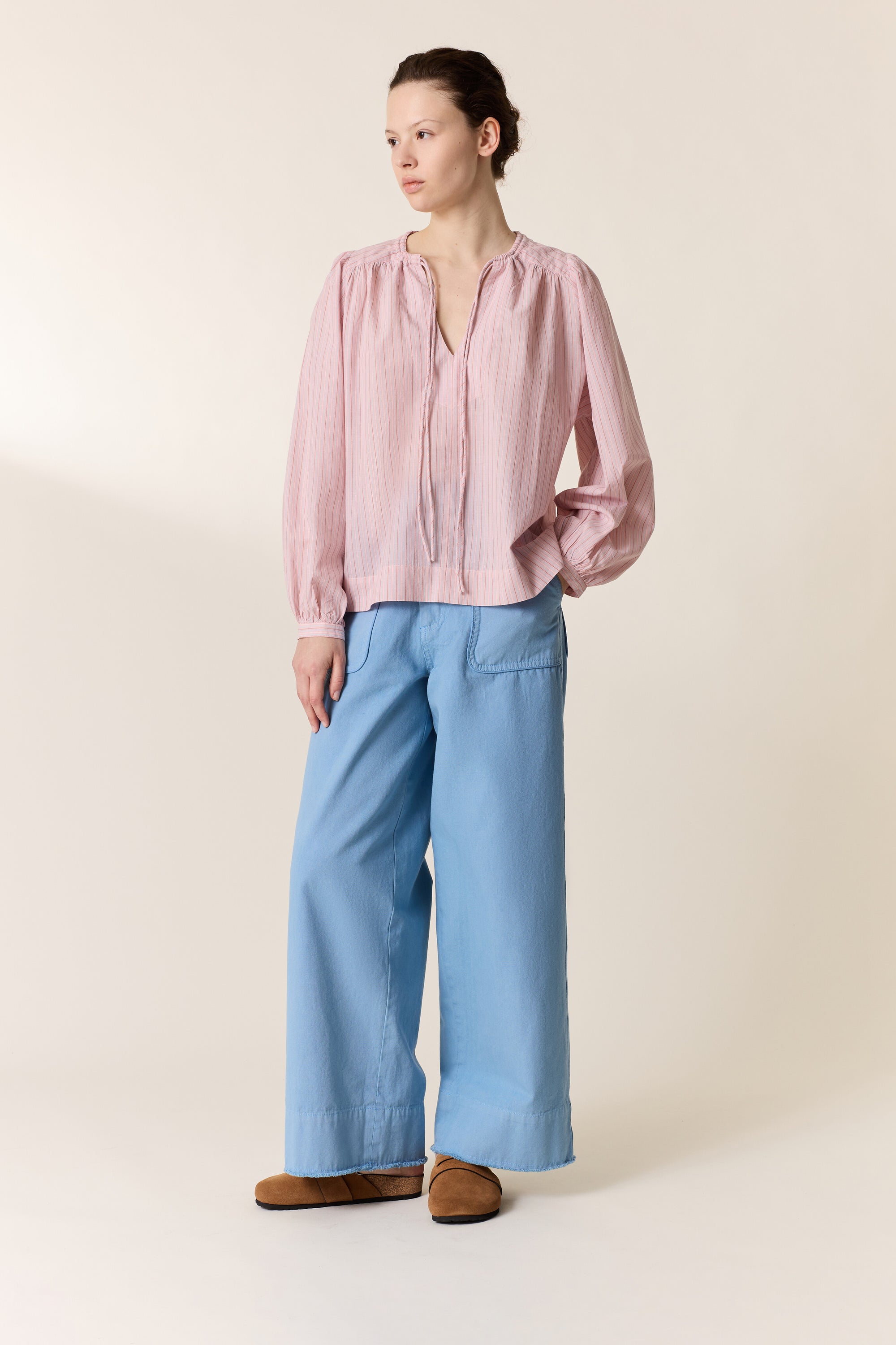 Woman wearing a pink stripe shirt  with light blue wide leg pants on a plain background. 