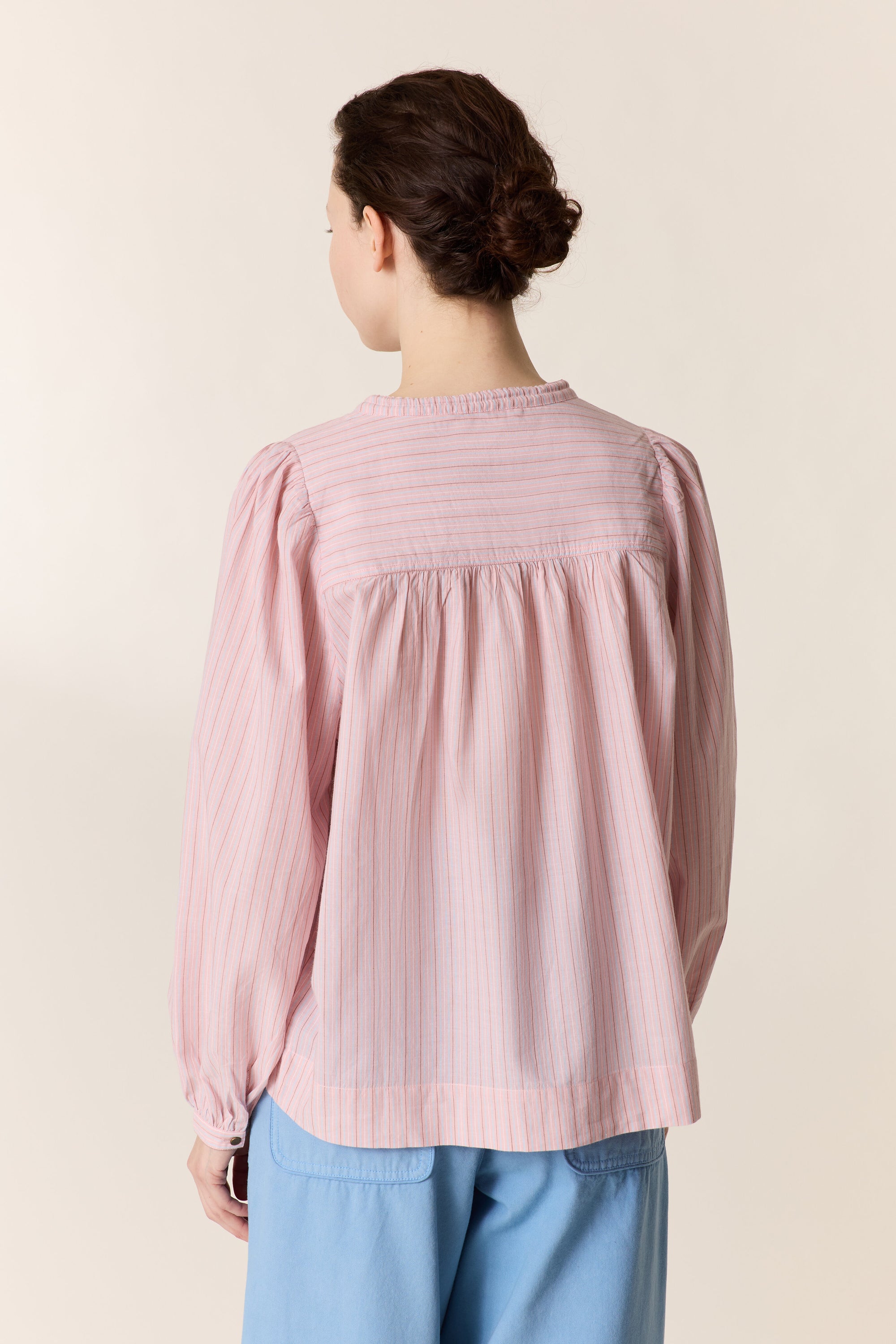 Woman shown from the back wearing a pink stripe shirt with light blue pants on a plain background. 