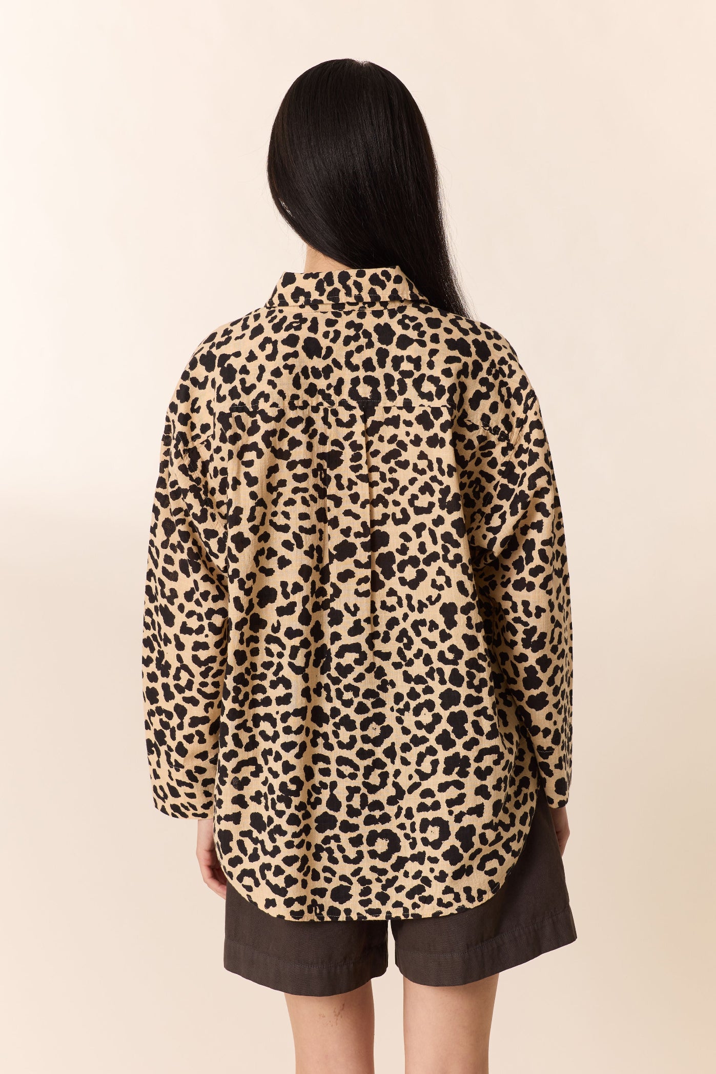 Woman shown from the back wearing a leopard print  shirt with brown shorts on a plain studio background. 