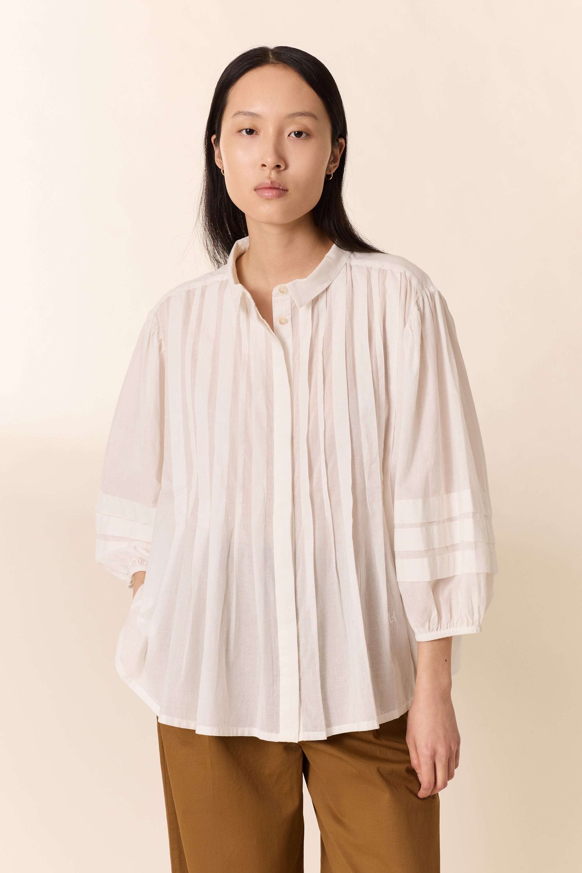 Women shown wearing a white pleated shirt  with tan wide pants on a plain background. 