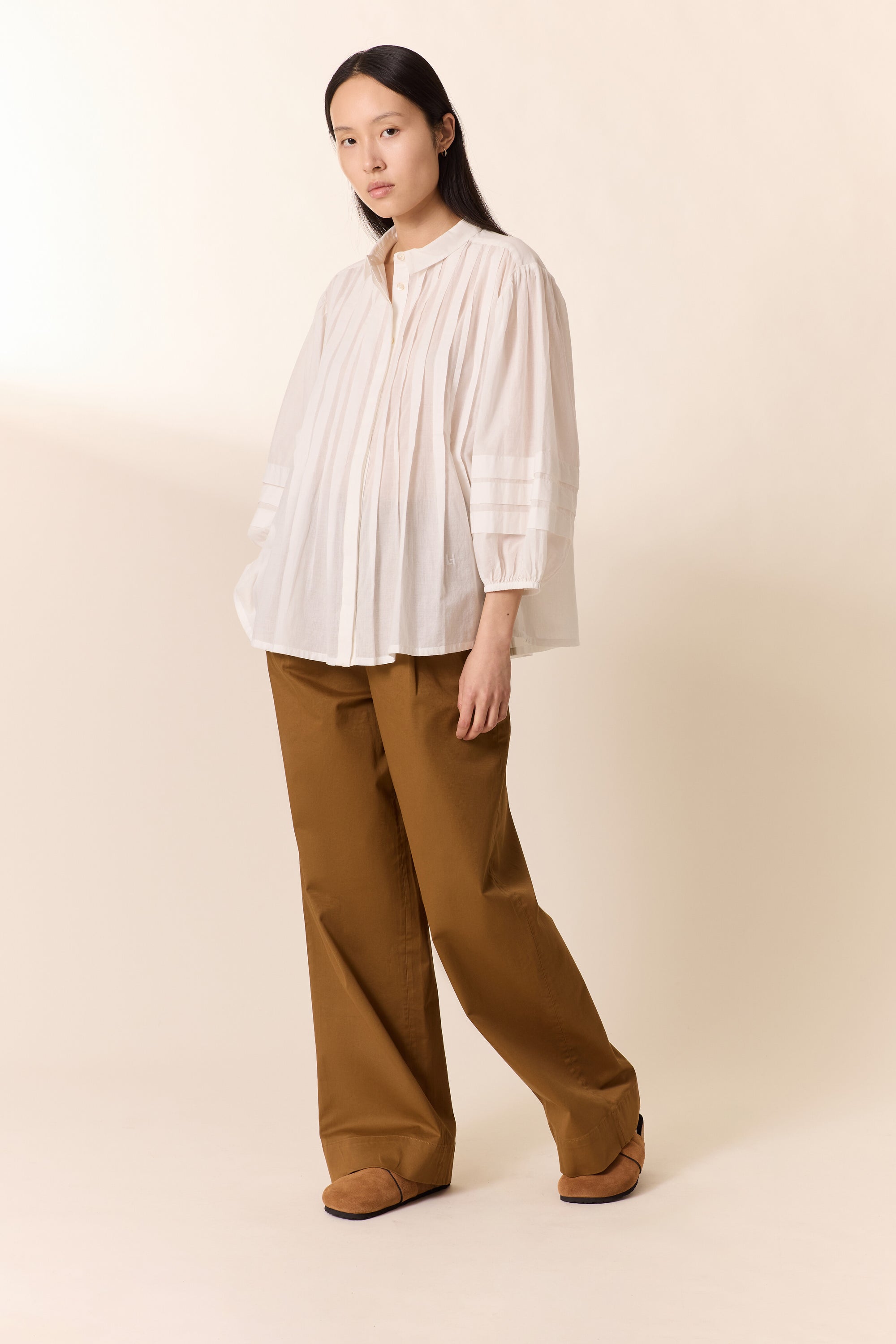 Women shown wearing a white pleated shirt  with tan wide pants on a plain background. 