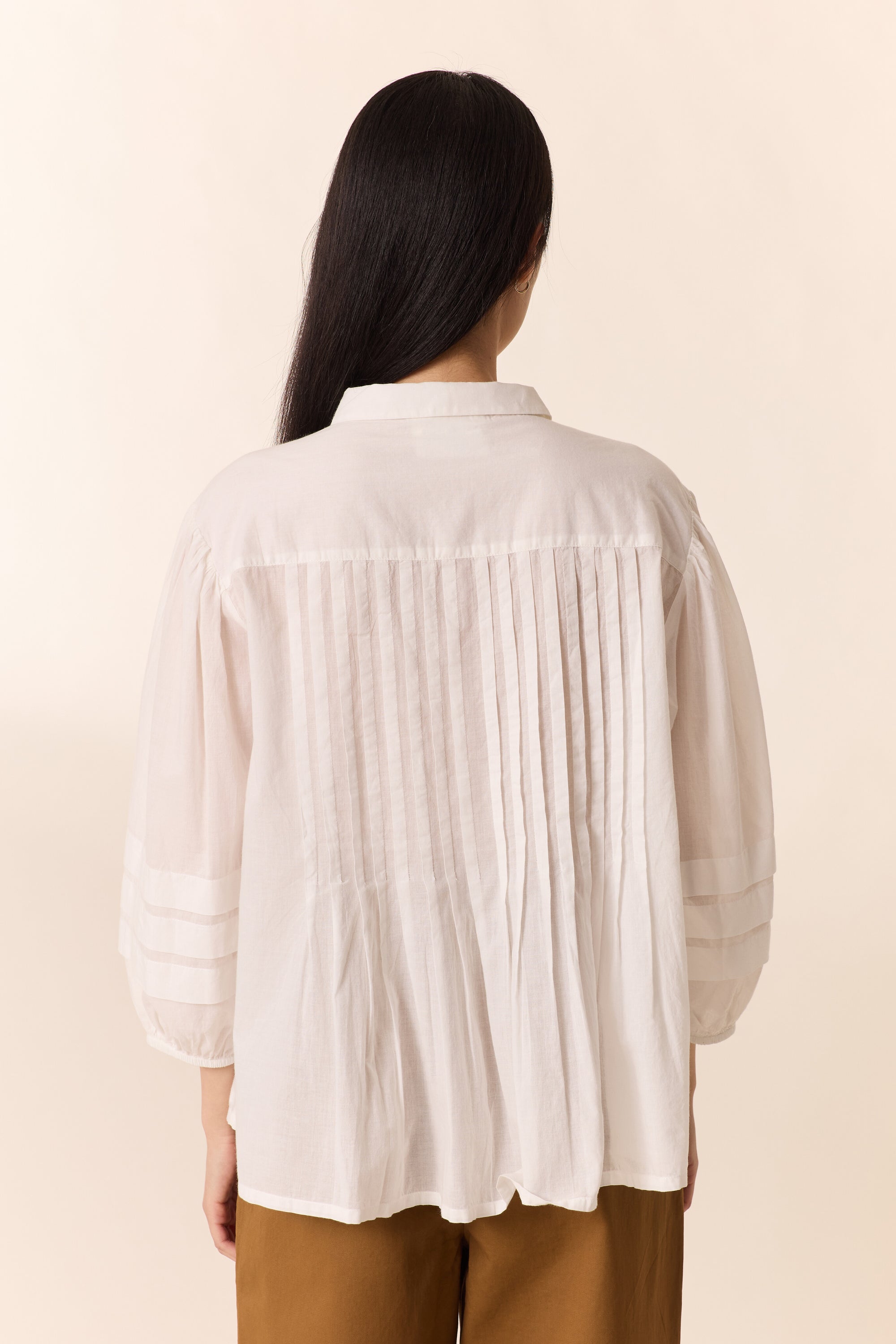 Women shown from the back with white pleated shirt on a plain background. 
