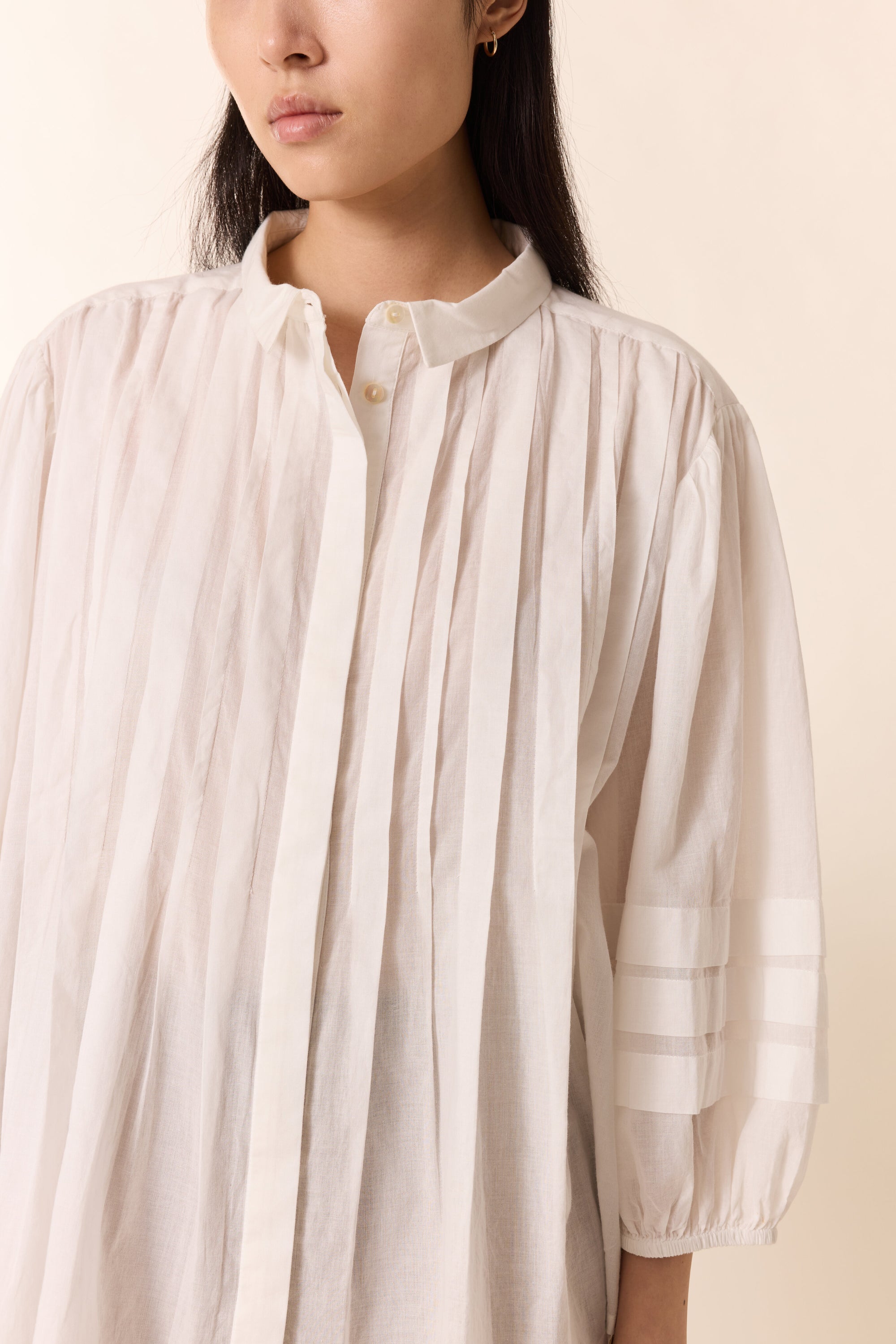 Women shown close up with white pleated shirt on a plain background. 