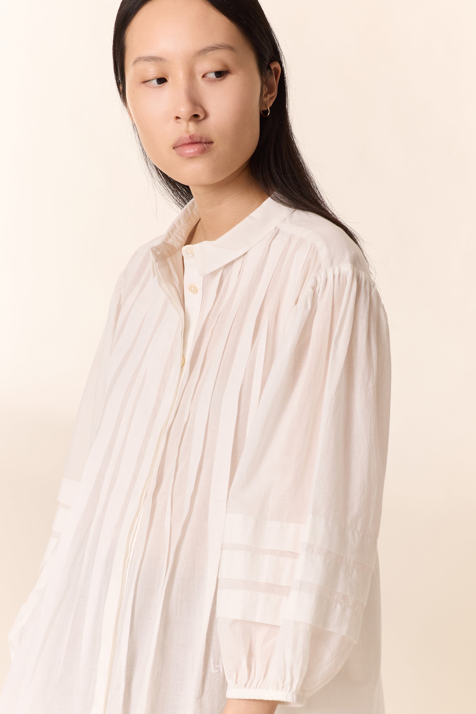 Women shown close up with white pleated shirt on a plain background. 