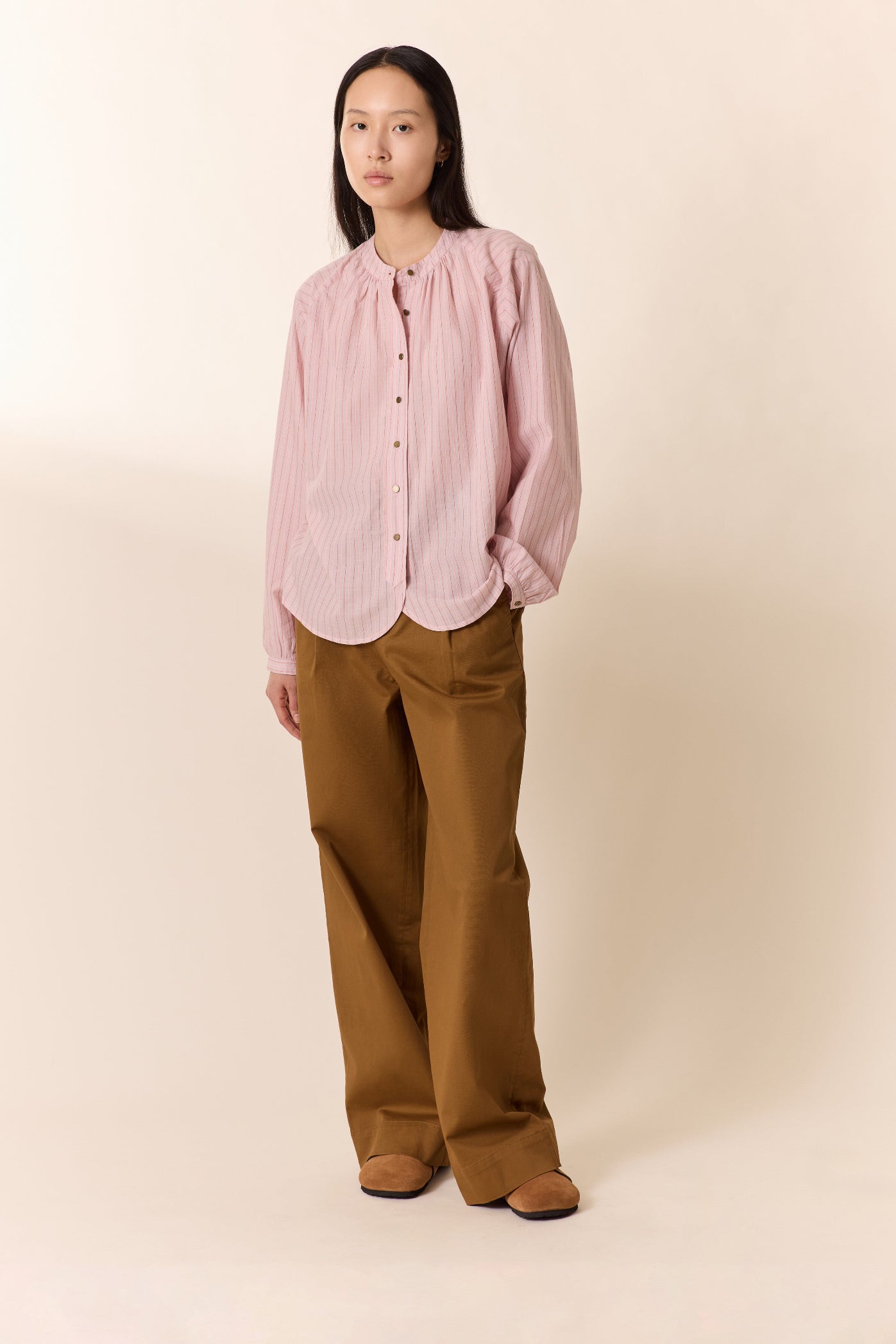 A woman stands against a plain beige studio background wearing a light pink vertically striped button-up blouse with a high gathered neckline and wide-leg brown pants.