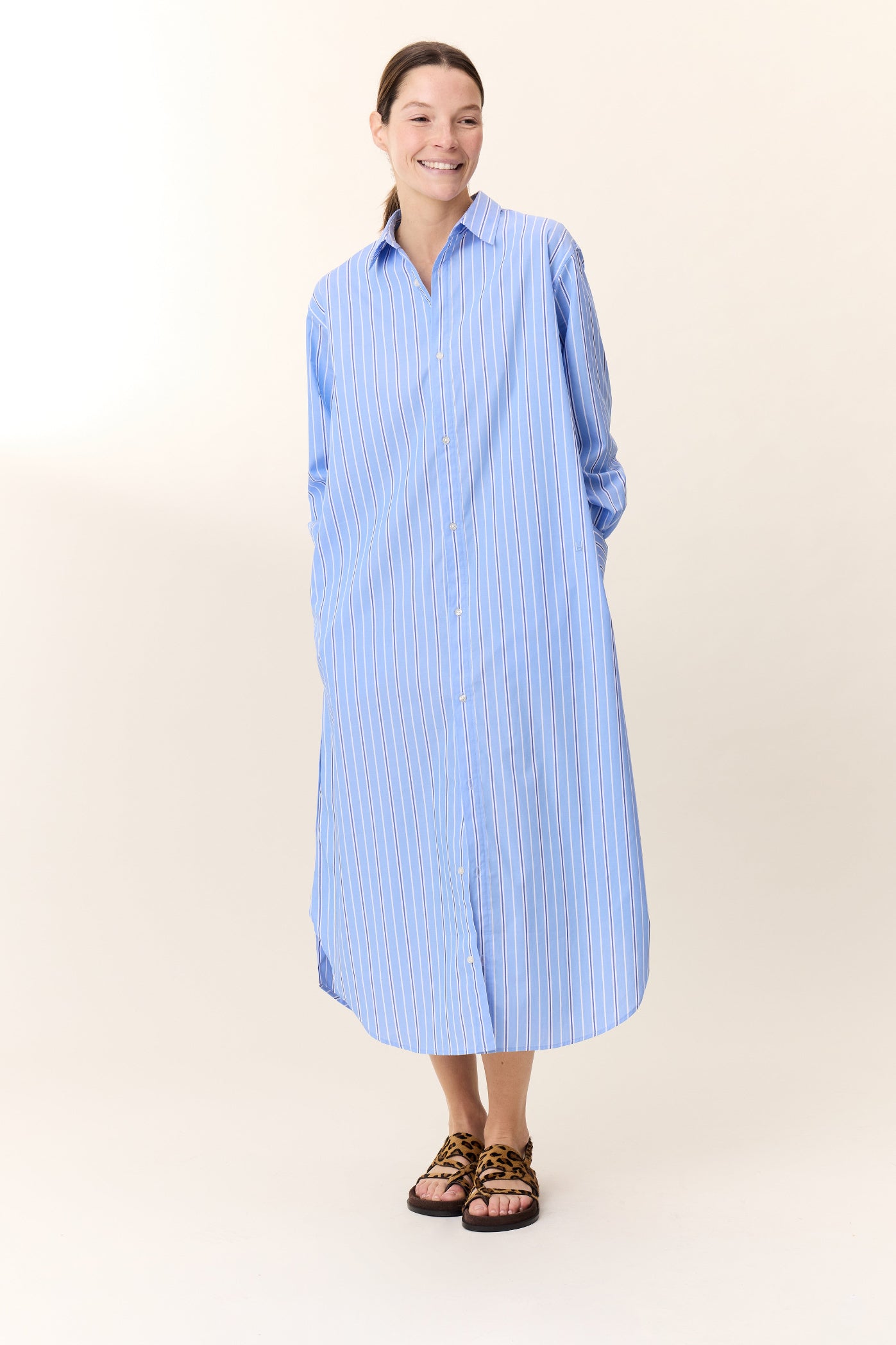 Model wearing long sleeve blue striped shirt dress with leopard print sandals on a white background. 