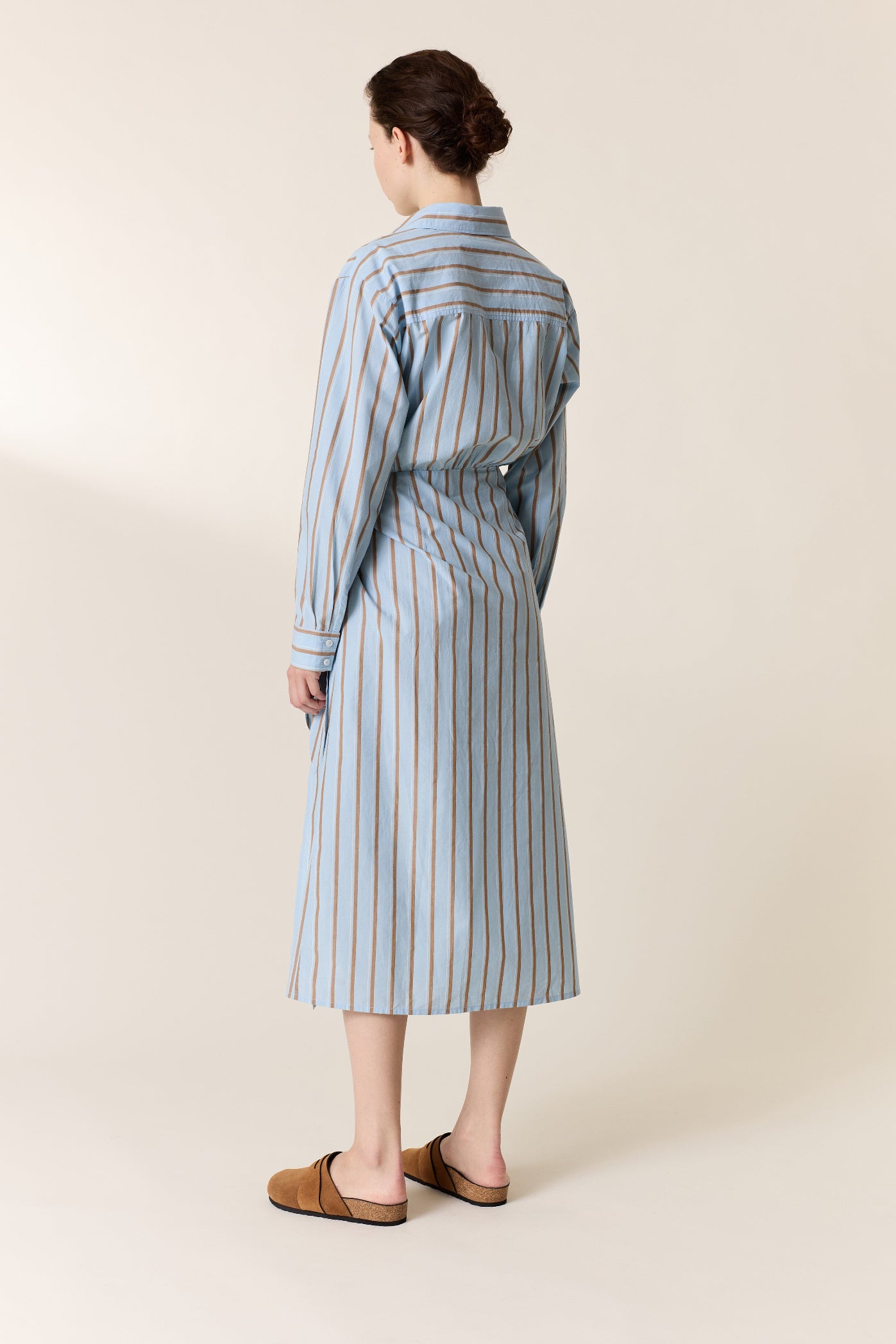 A woman stands with her back to the camera wearing a light blue and white vertically striped shirt dress with brown mule sandals against a warm beige studio background.