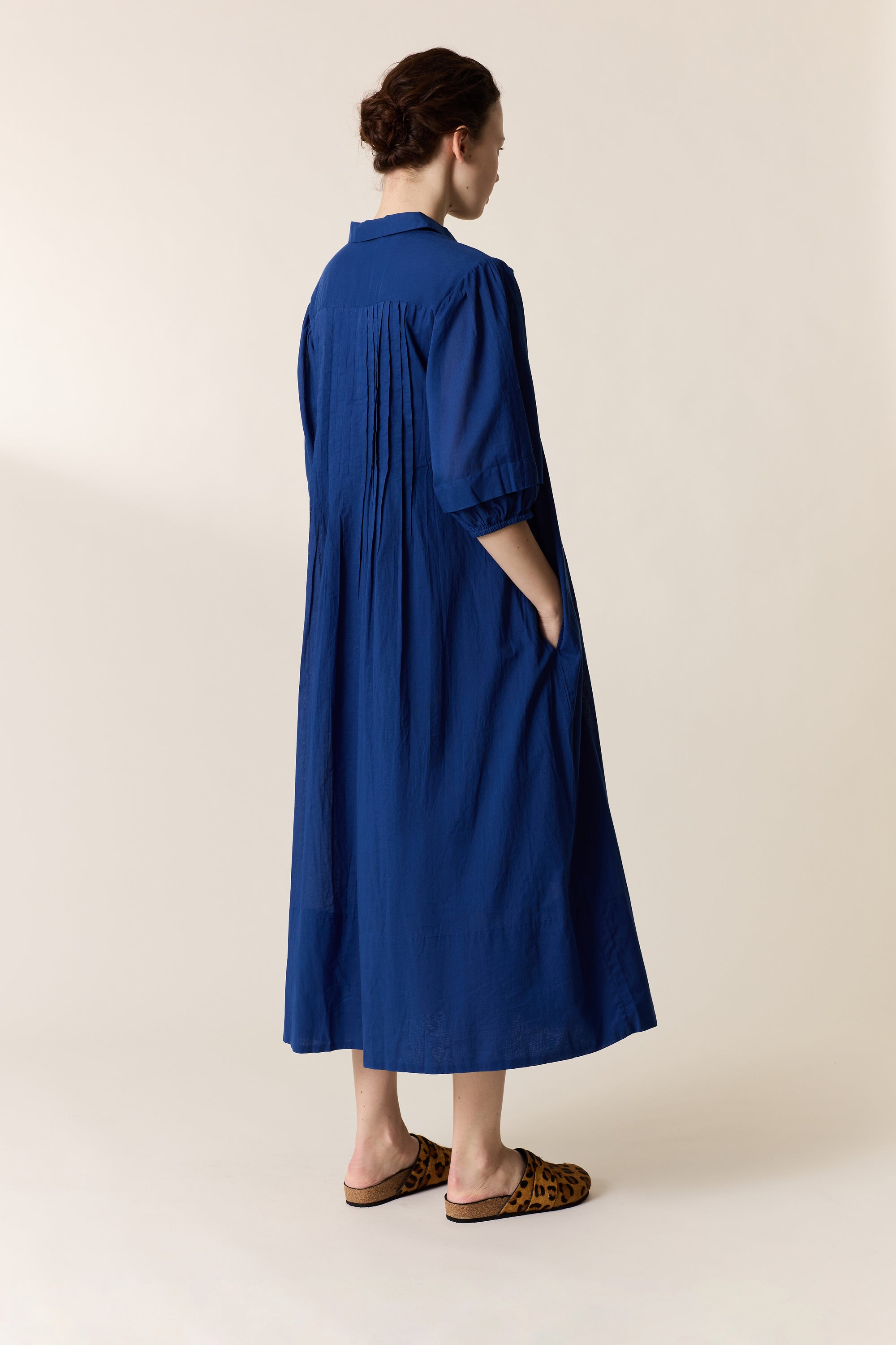 Women shown from the back n wearing a blue pleated dress on a plain background
