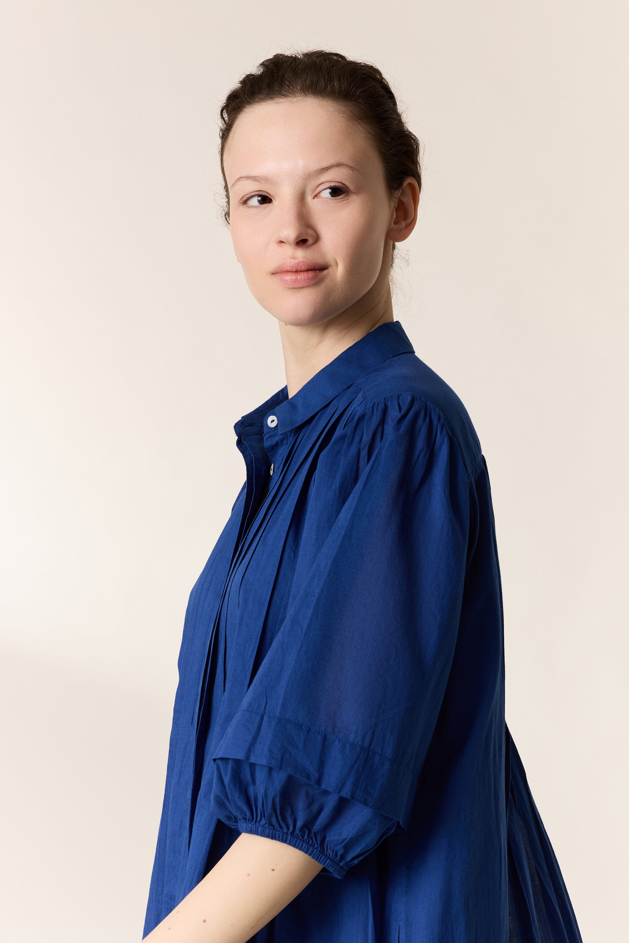 Women shown side on wearing a blue pleated dress on a plain background. 