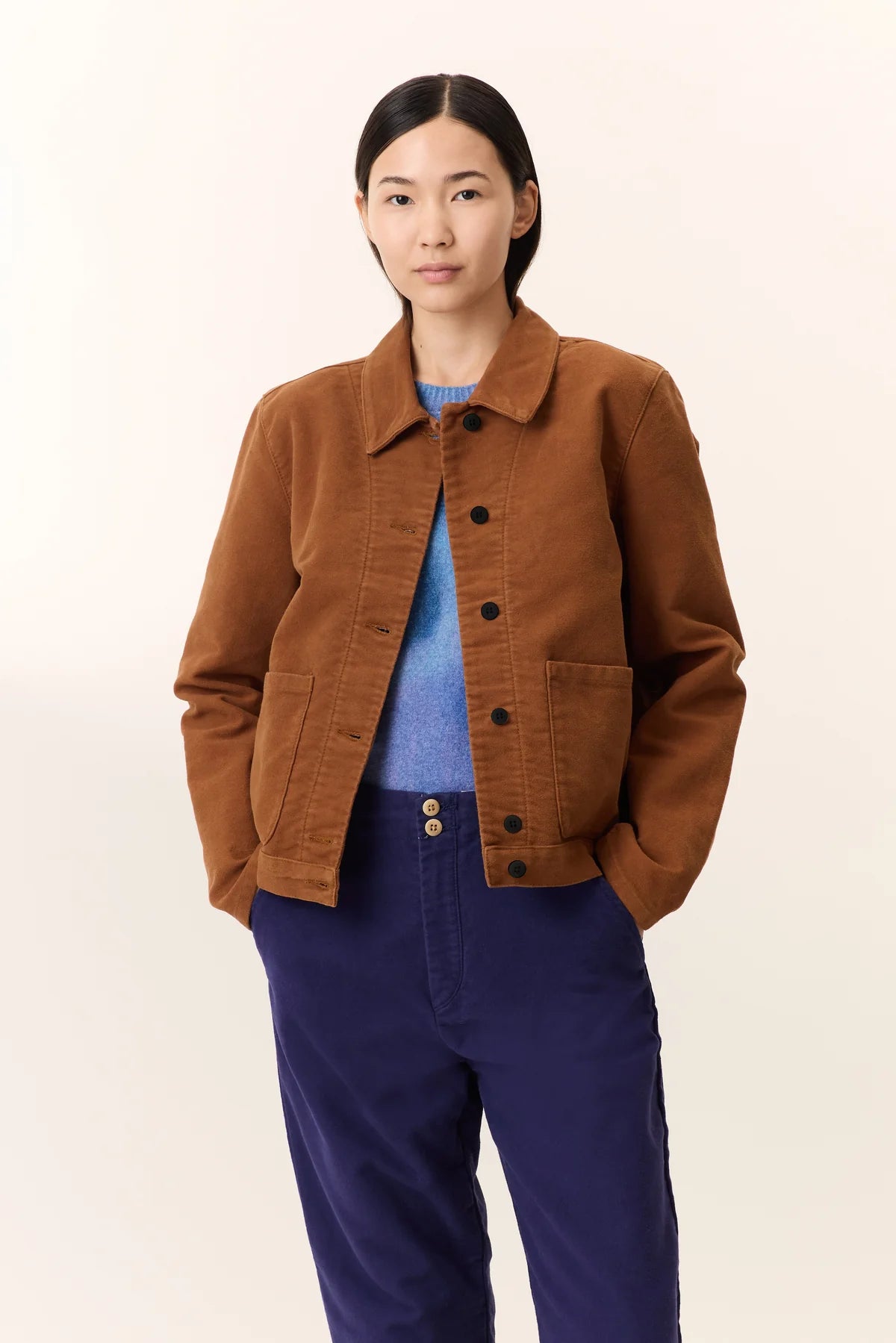 Person wearing a camel brown jacket with blue jumper under and blue pants on a white background