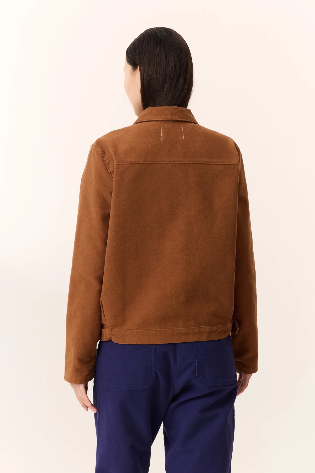 Person wearing a camel brown jacket and blue pants on a beige background