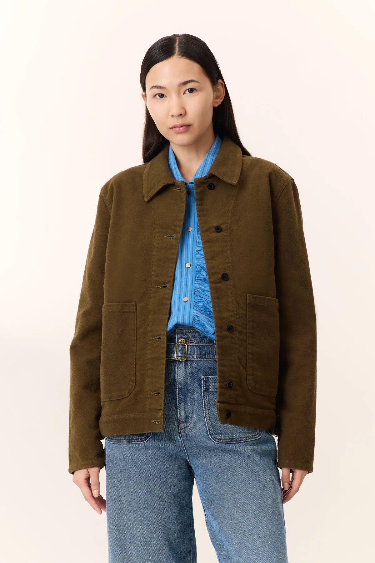 Person wearing a khaki jacket with a blue shirt underneath and blue jeans on a beige background