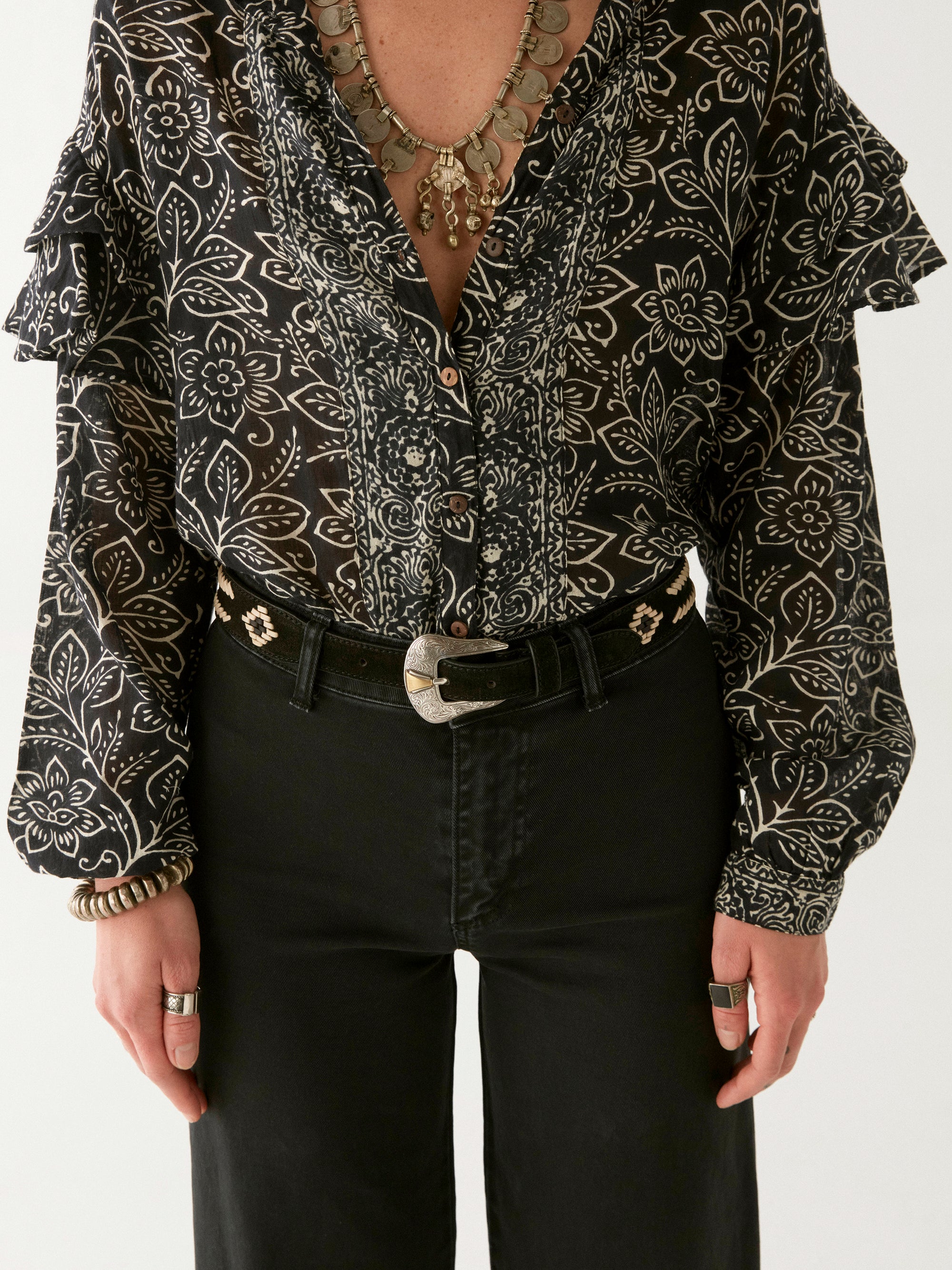 Woman wearing a black and ivory floral patterned blouse up close to show button detail and frill sleeve. Paired with black pants and a belt.  