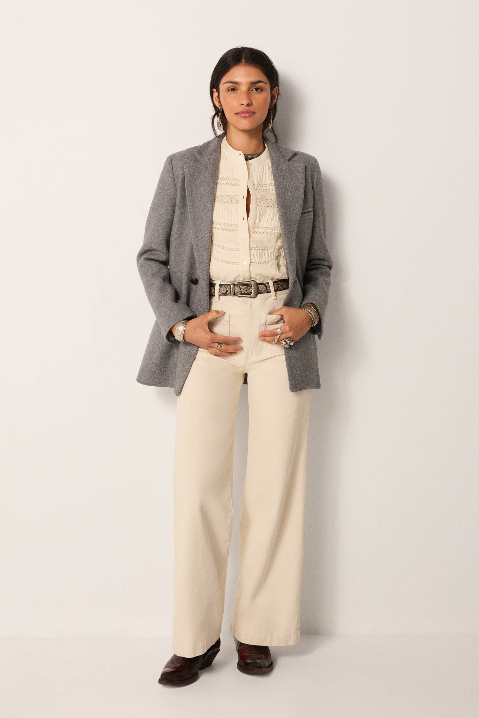 Woman wearing a gray blazer over a cream outfit with white pants on a plain background