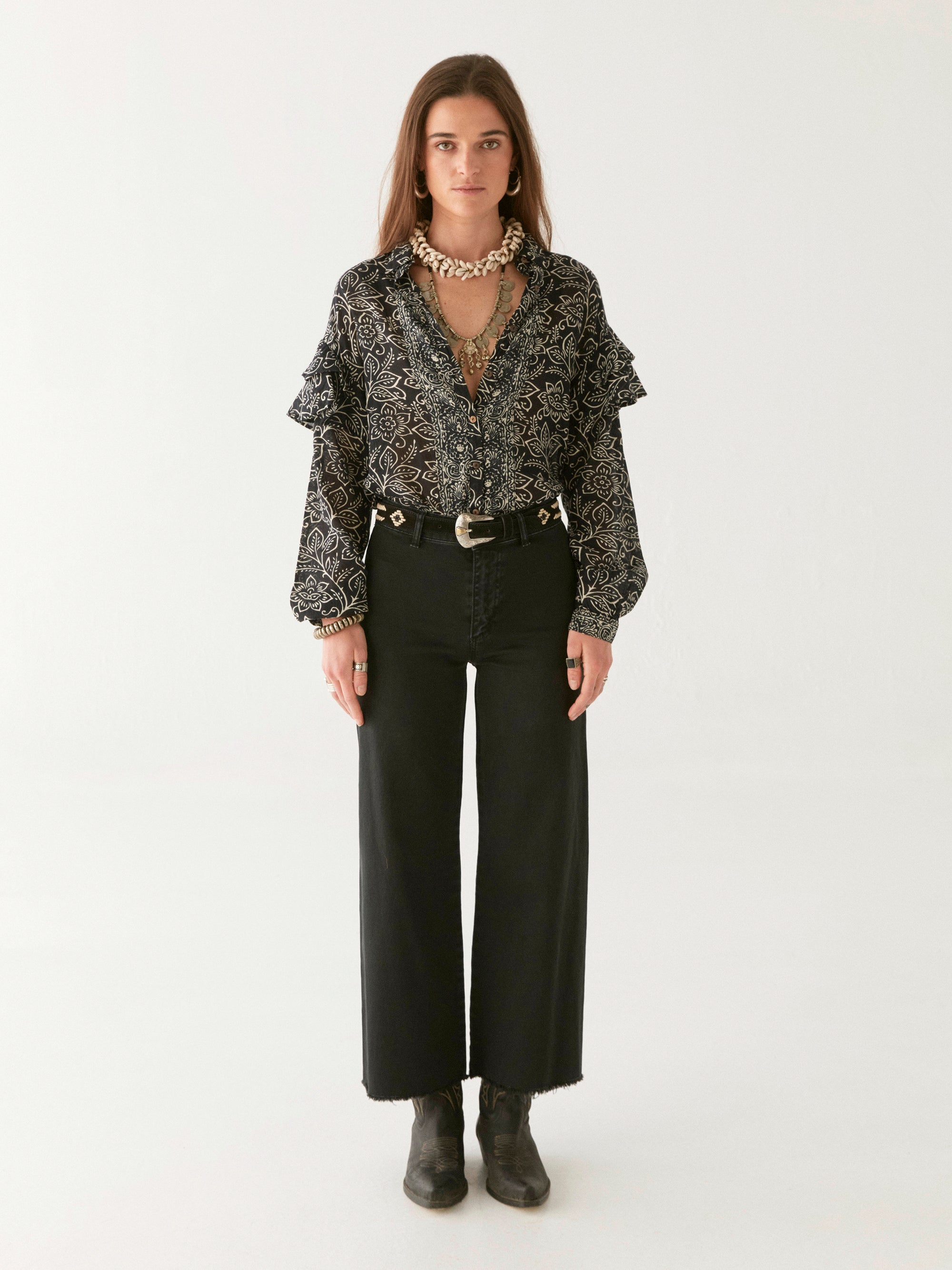 Woman wearing a black and ivory floral patterned blouse, black pants, and boots on a white background. 