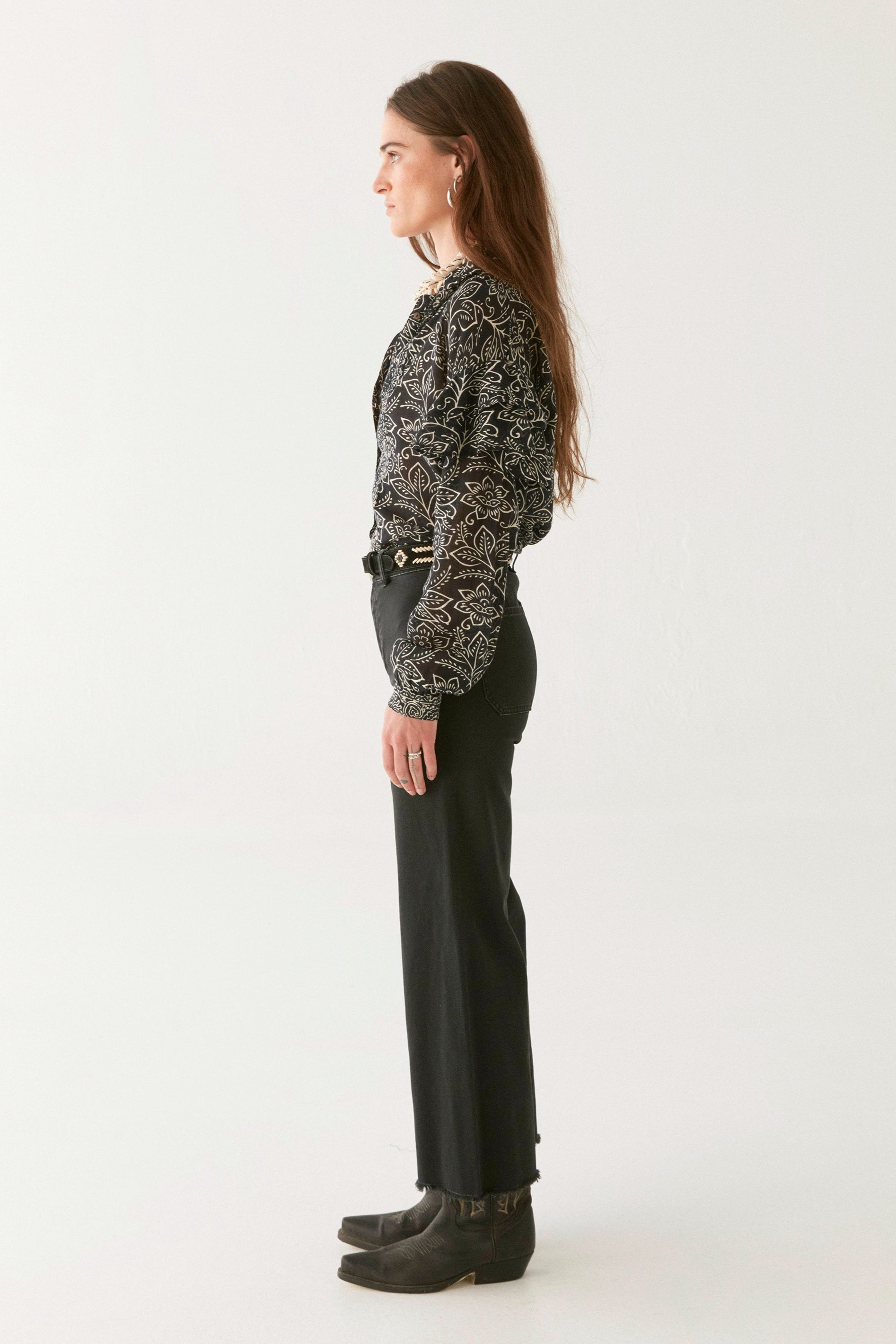 Woman wearing a black and ivory floral patterned blouse, black pants, and boots on a white background. 