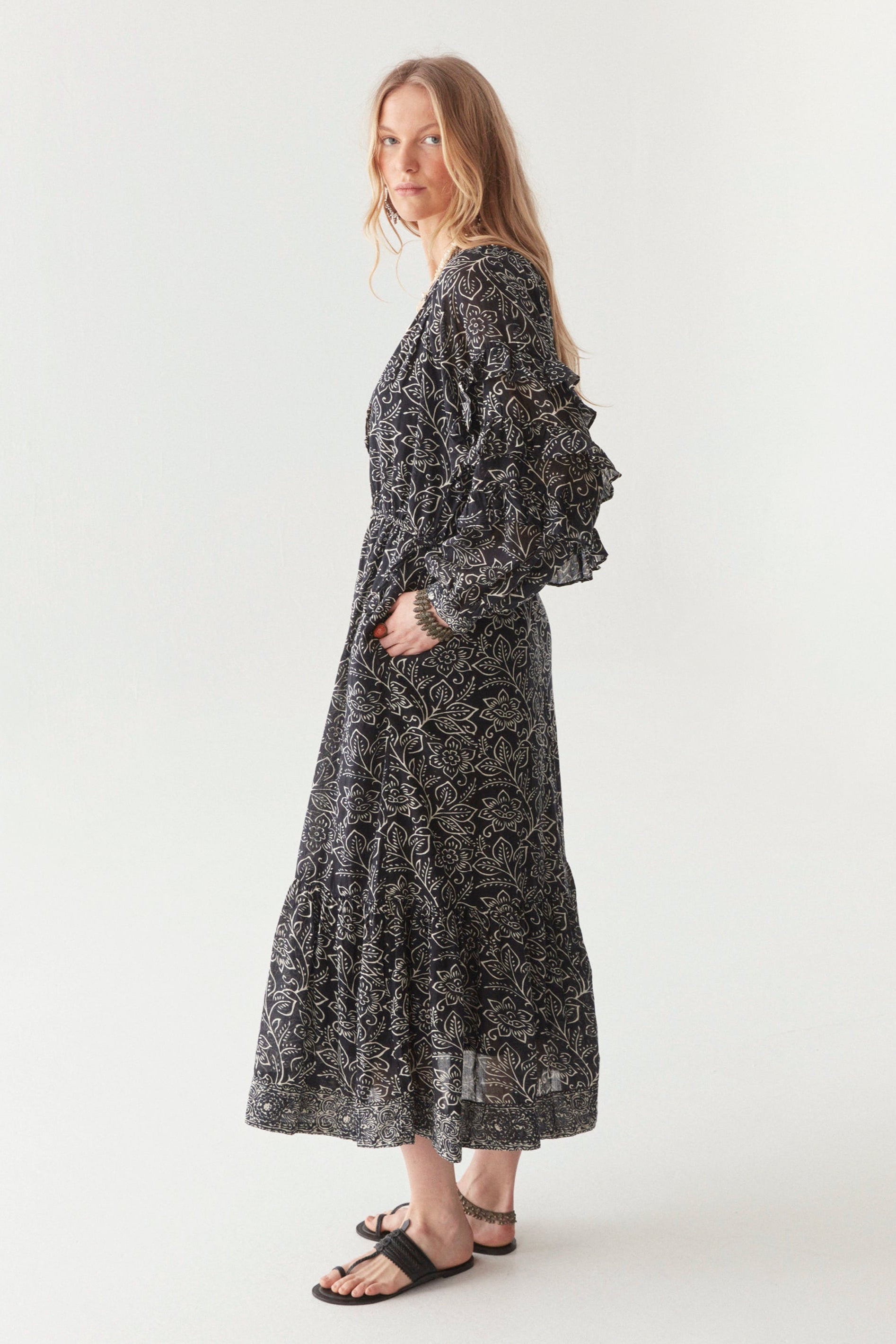 Woman wearing a black and white floral patterned maxi dress against a white background. Showing frill detail on the long sleeves. 