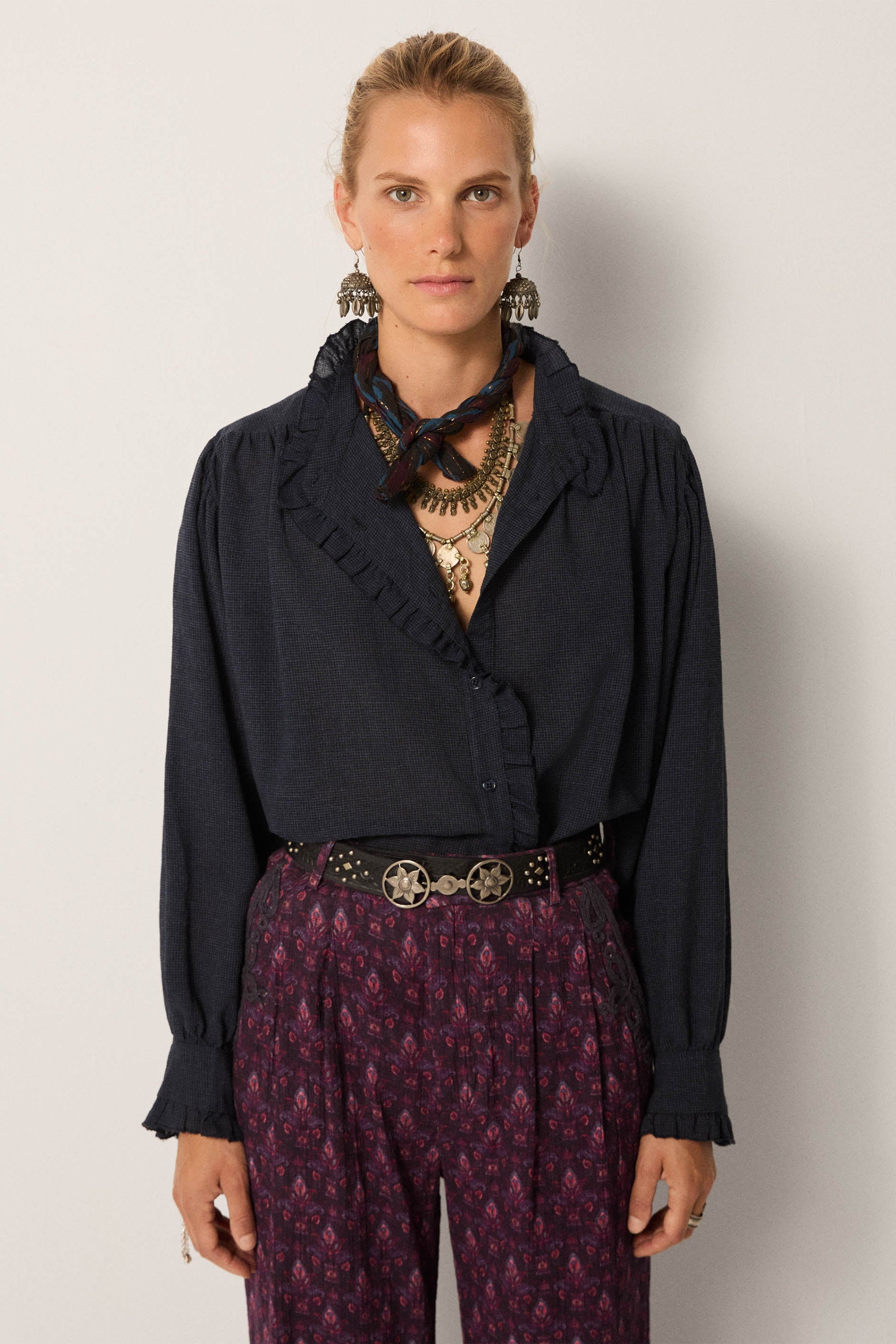 Woman wearing a dark blue blouse with ruffled details and patterned purple pants against a plain background