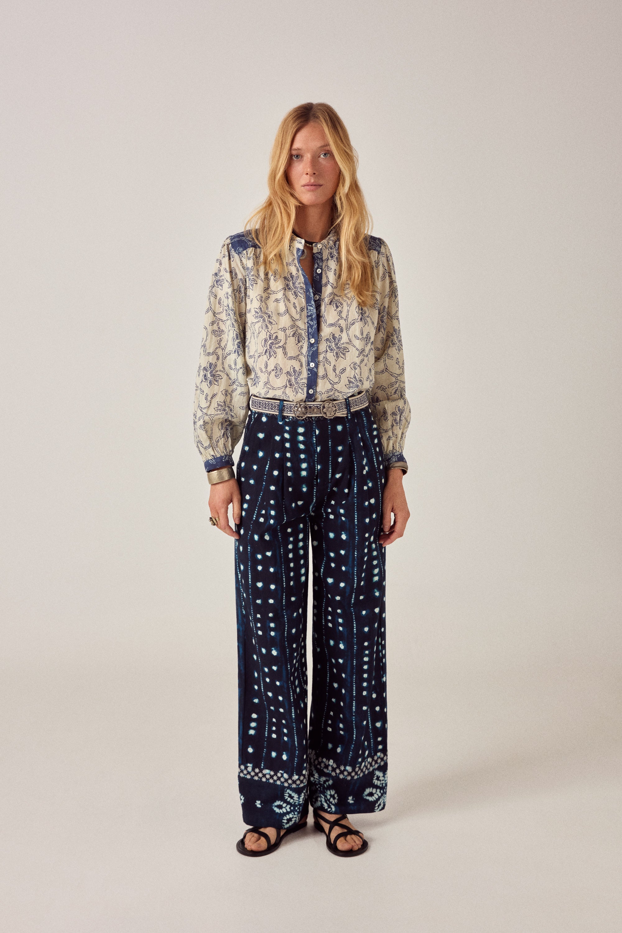 Woman wearing a patterned blouse and navy pants with white patterns on a light background