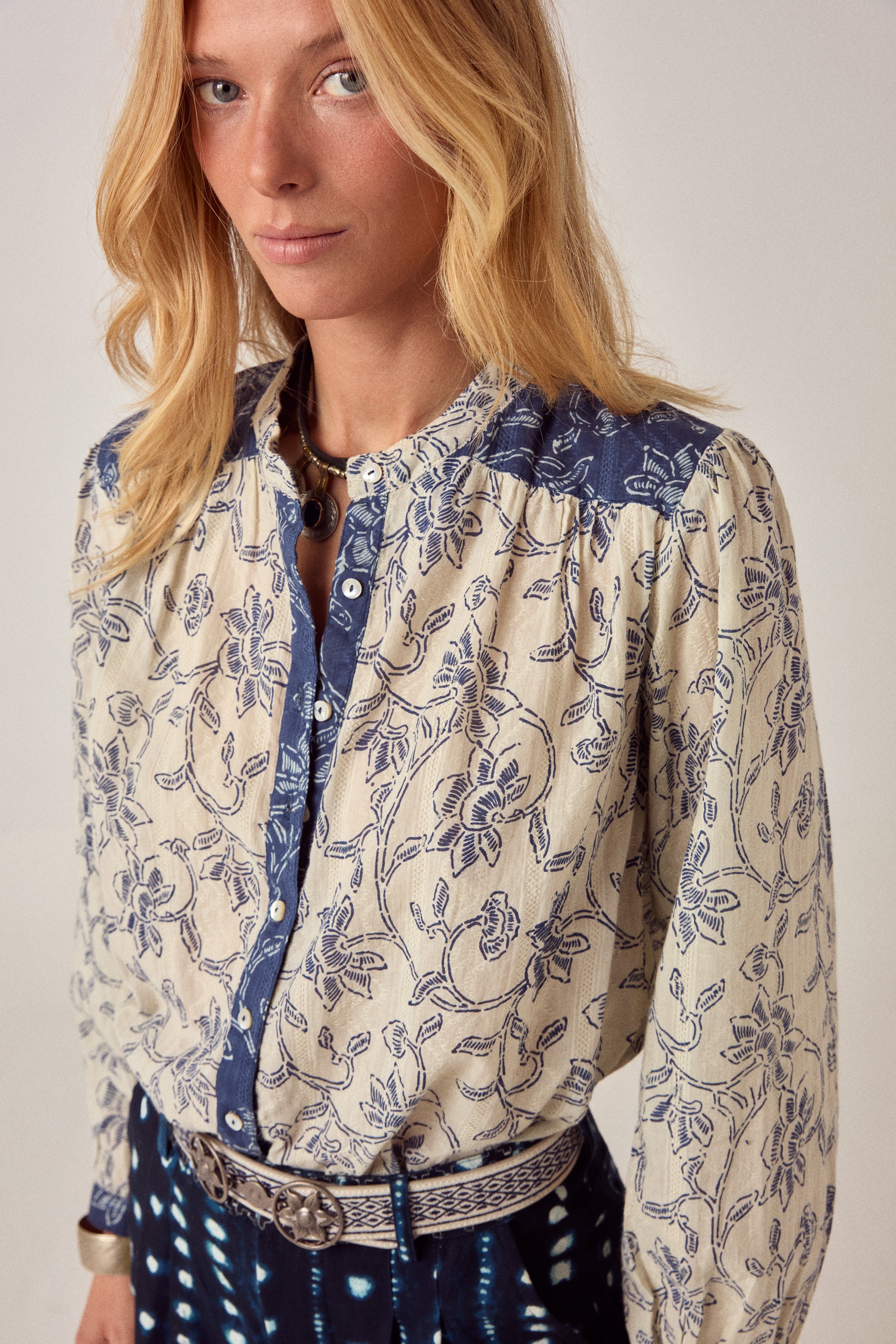 Woman wearing a patterned blouse and navy pants with white patterns on a light background
