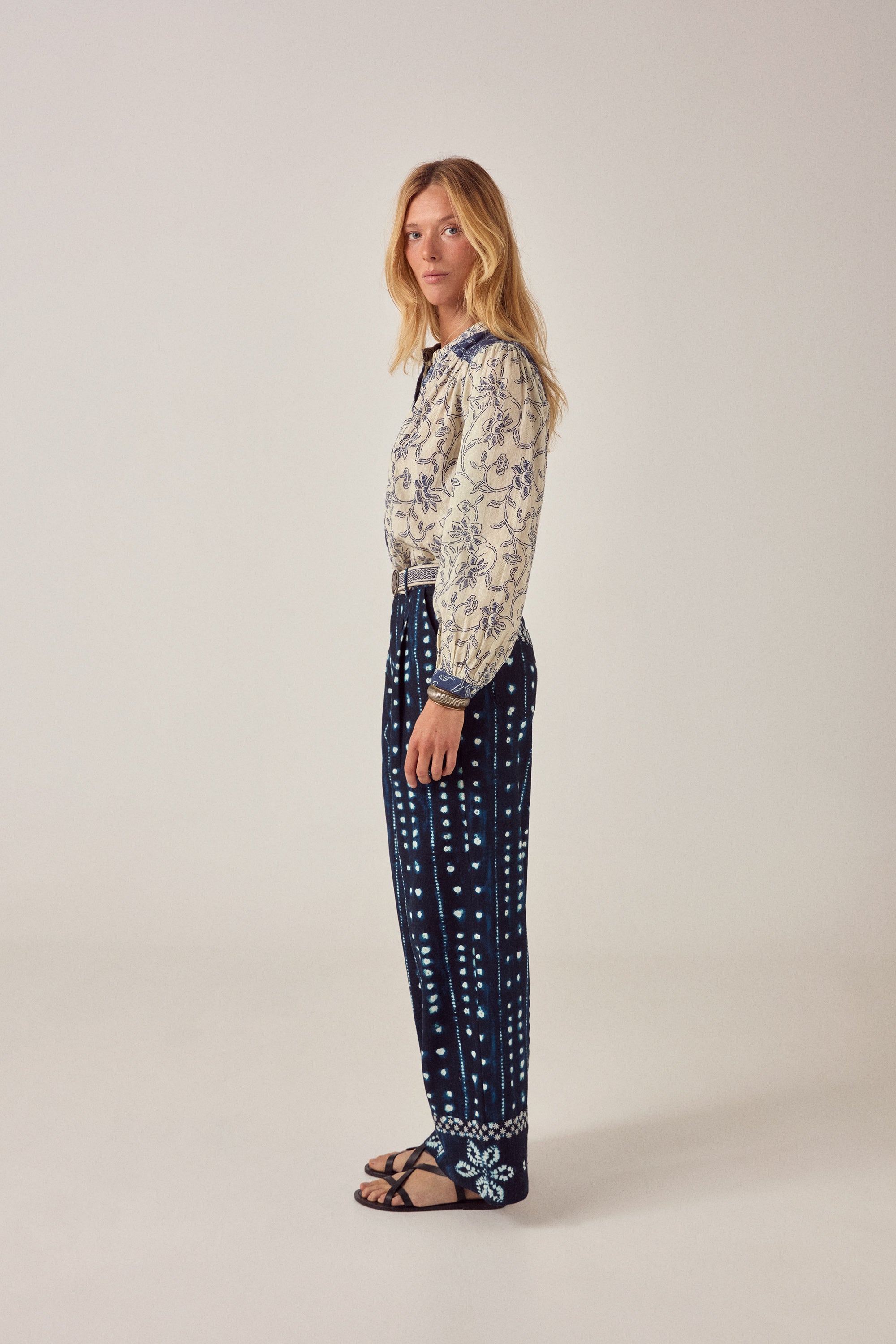 Woman wearing a patterned blouse and navy pants with white patterns on a light background