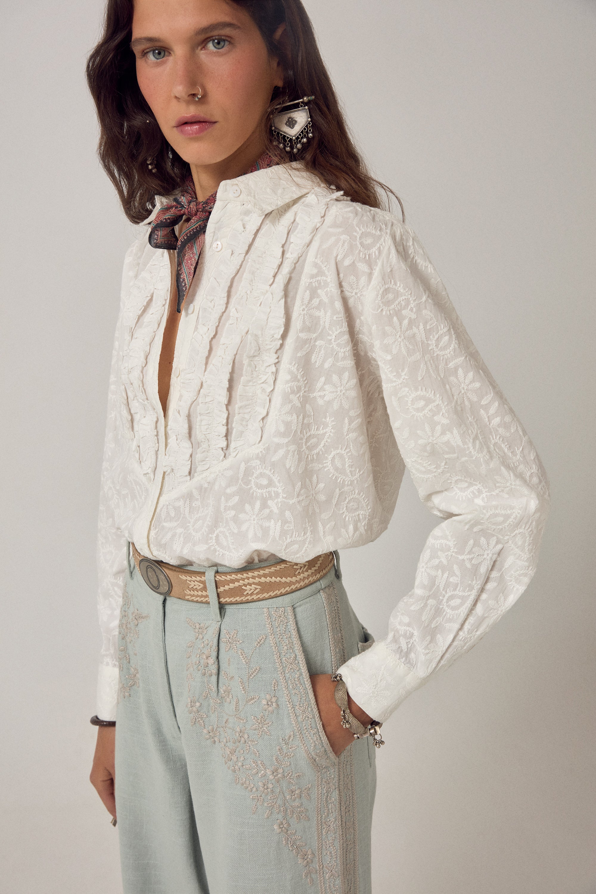 Woman wearing ecru long sleeve embroidered shirt with frill detail on a plain background. 