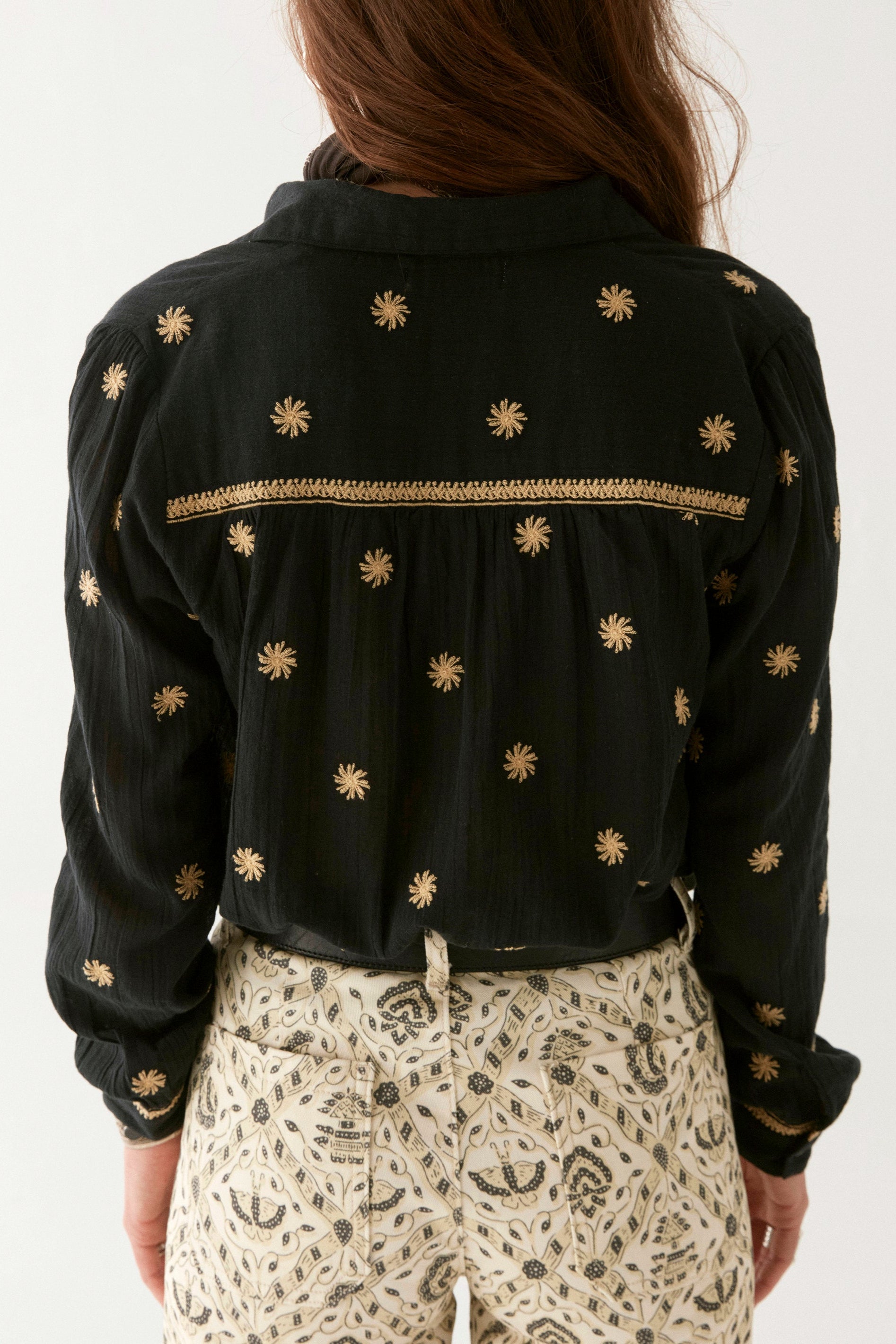 Model wearing black cotton blouse with gold embroidery. Paired with cream patterned trousers on a white background.  