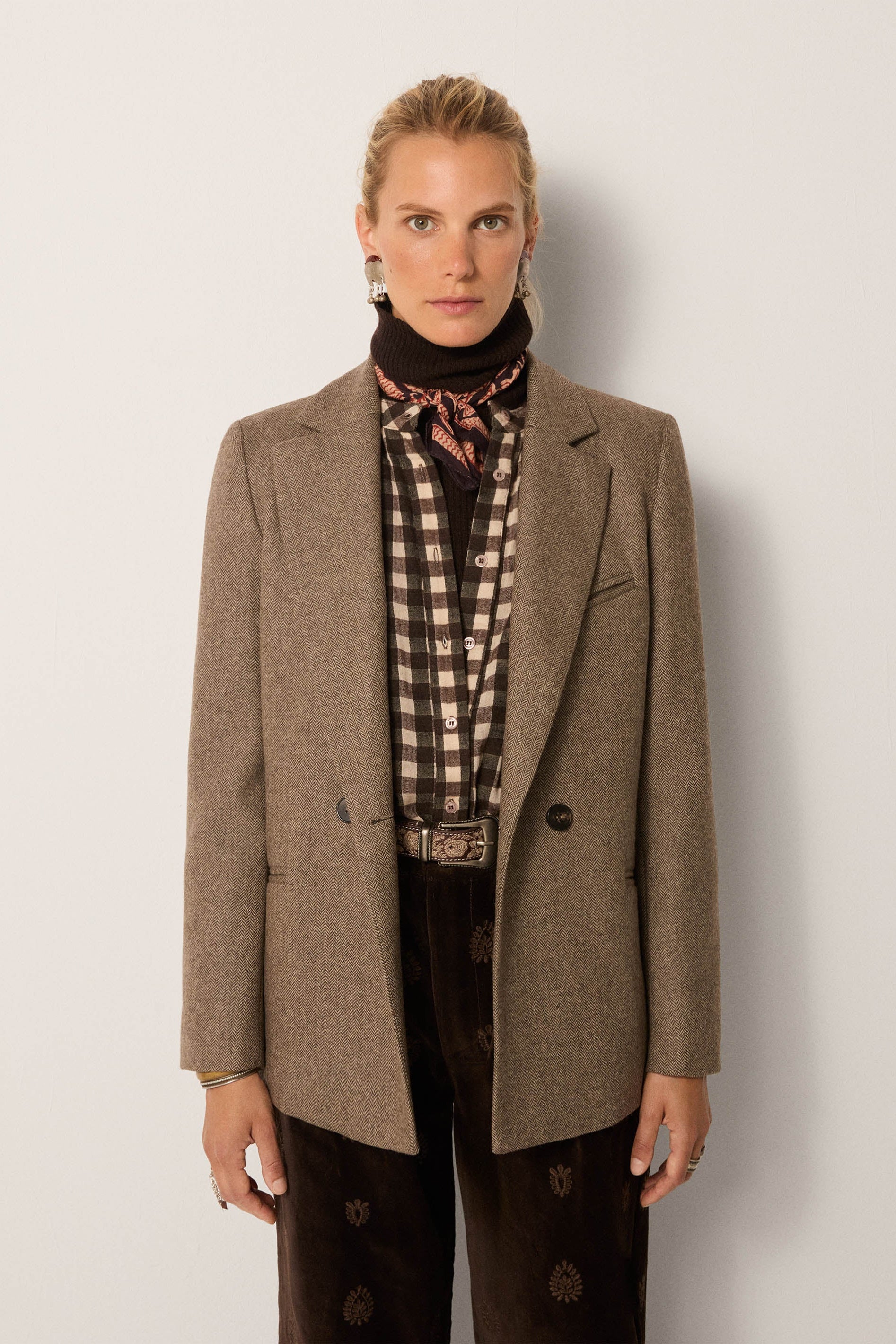 Person wearing a brown blazer over a plaid shirt with a patterned scarf, on a plain background