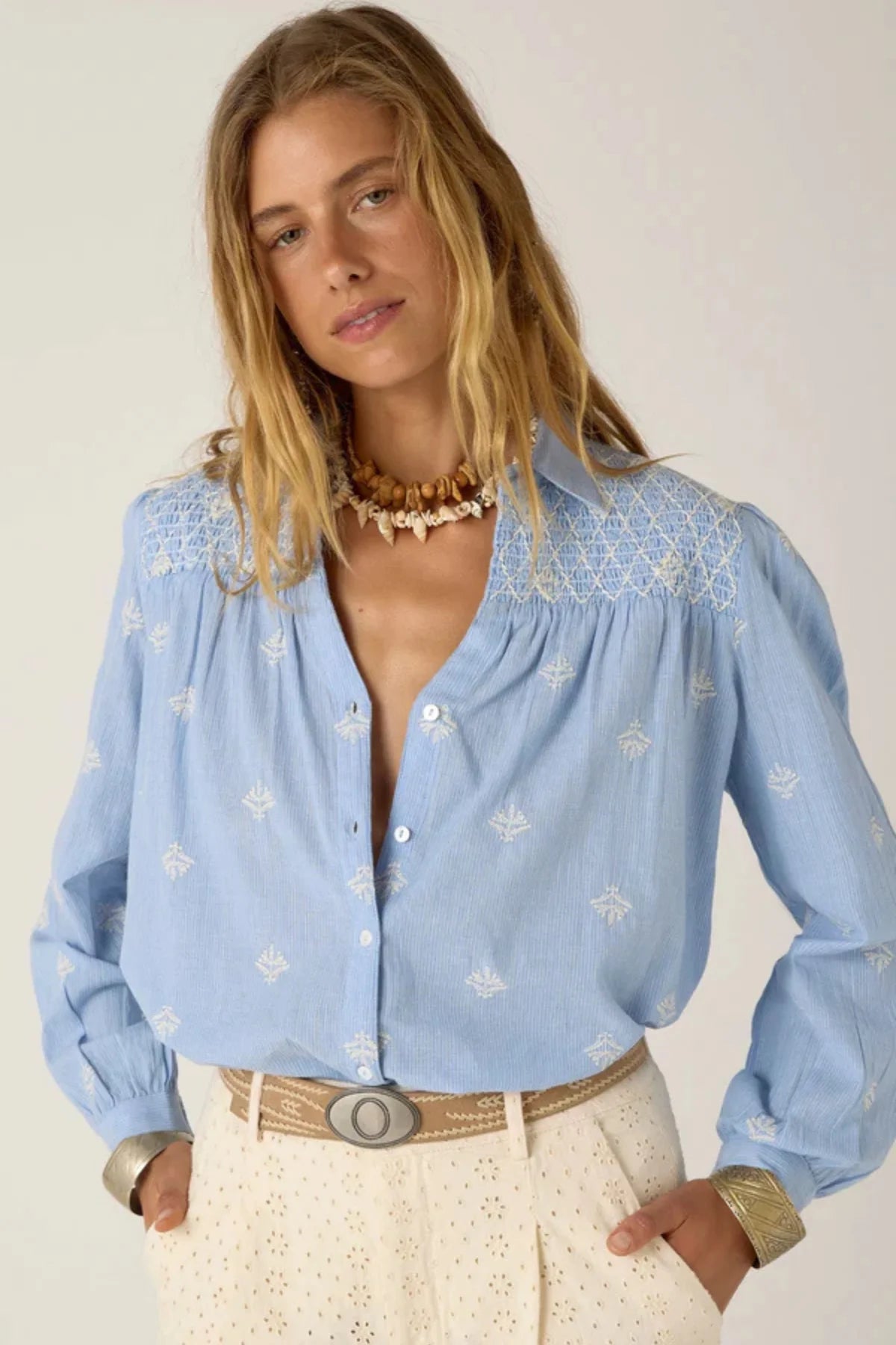 Women wearing a light blue long-sleeve shirt with white embroidery and white pants on a plain background. 