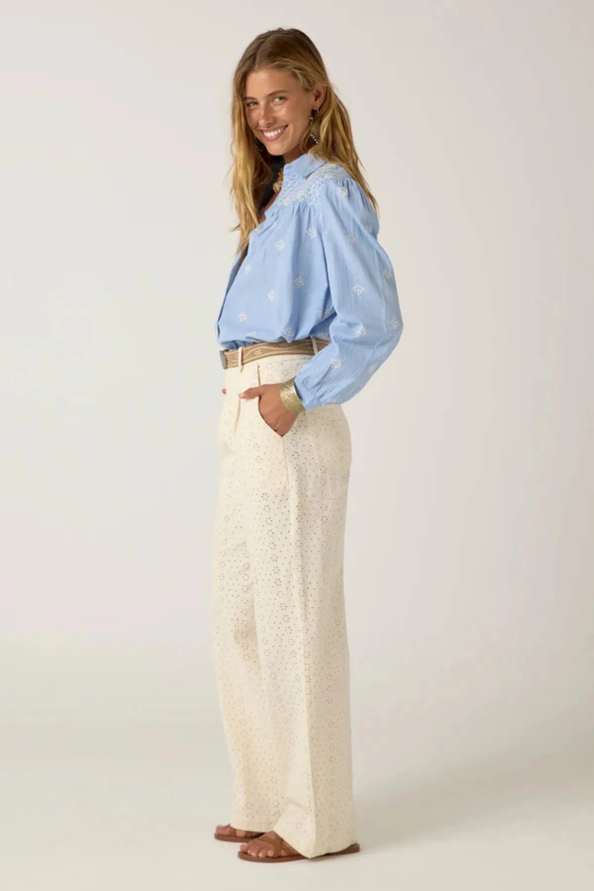 Women wearing a light blue long-sleeve shirt with white embroidery and white pants on a plain background. 