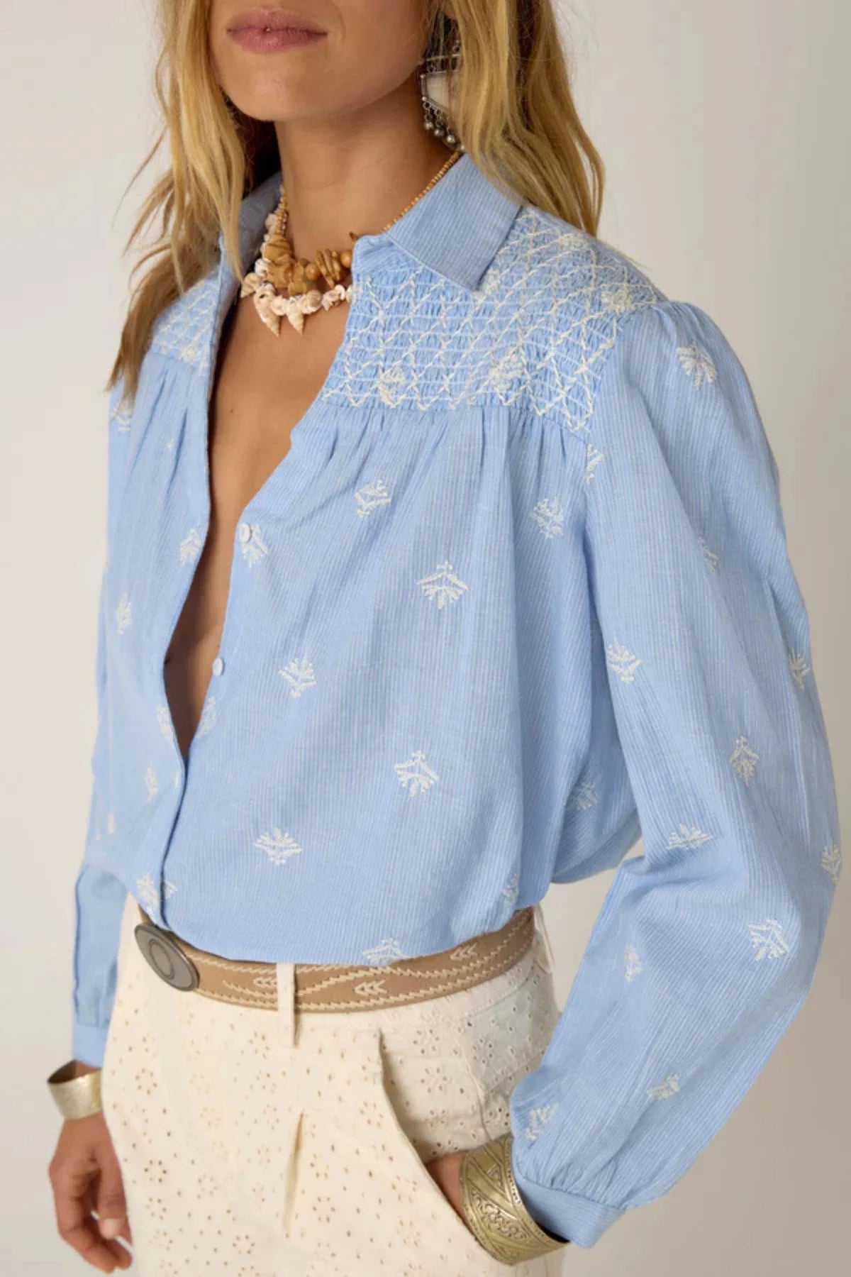 Women wearing a light blue long-sleeve shirt with white embroidery and white pants on a plain background. 