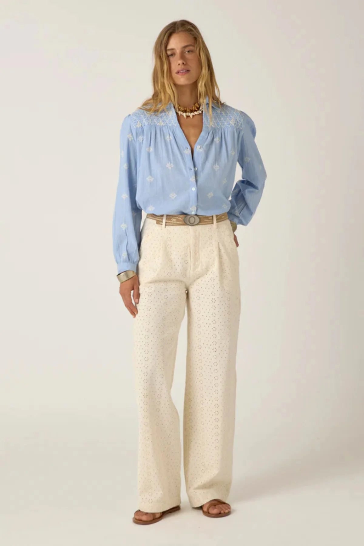 Women wearing a light blue long-sleeve shirt with white embroidery and white pants on a plain background. 