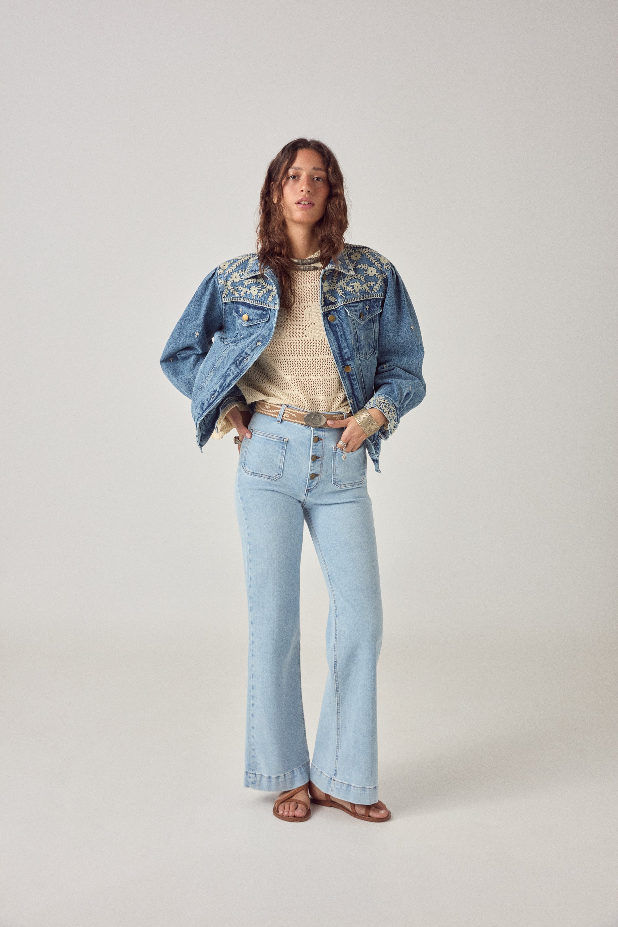 Woman wearing a blue denim jacket with white floral embroidery detailing with denim jeans on a plain background